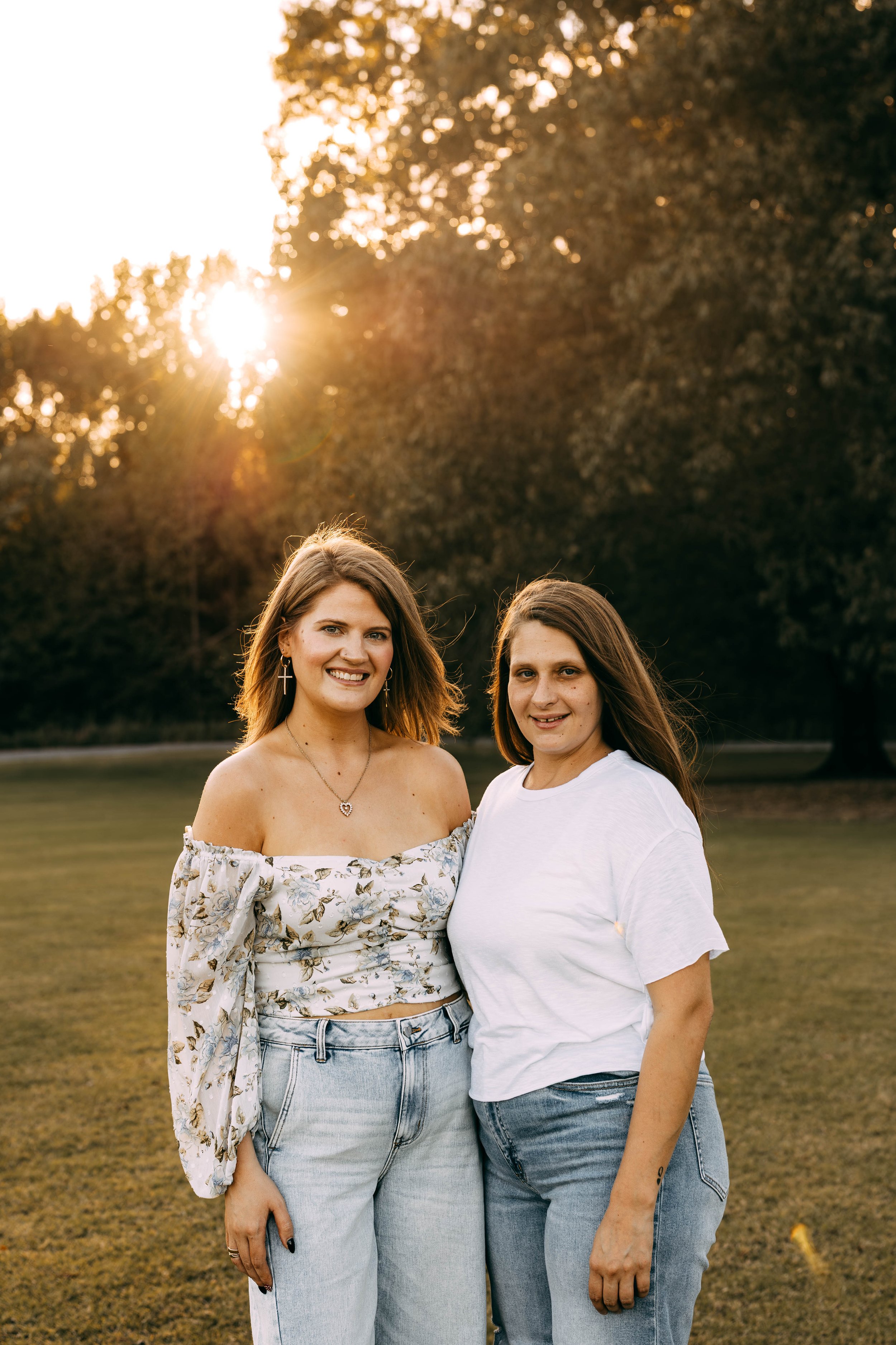 Two women standing outdoors at sunset, smiling, with trees and golden sunlight in the background.