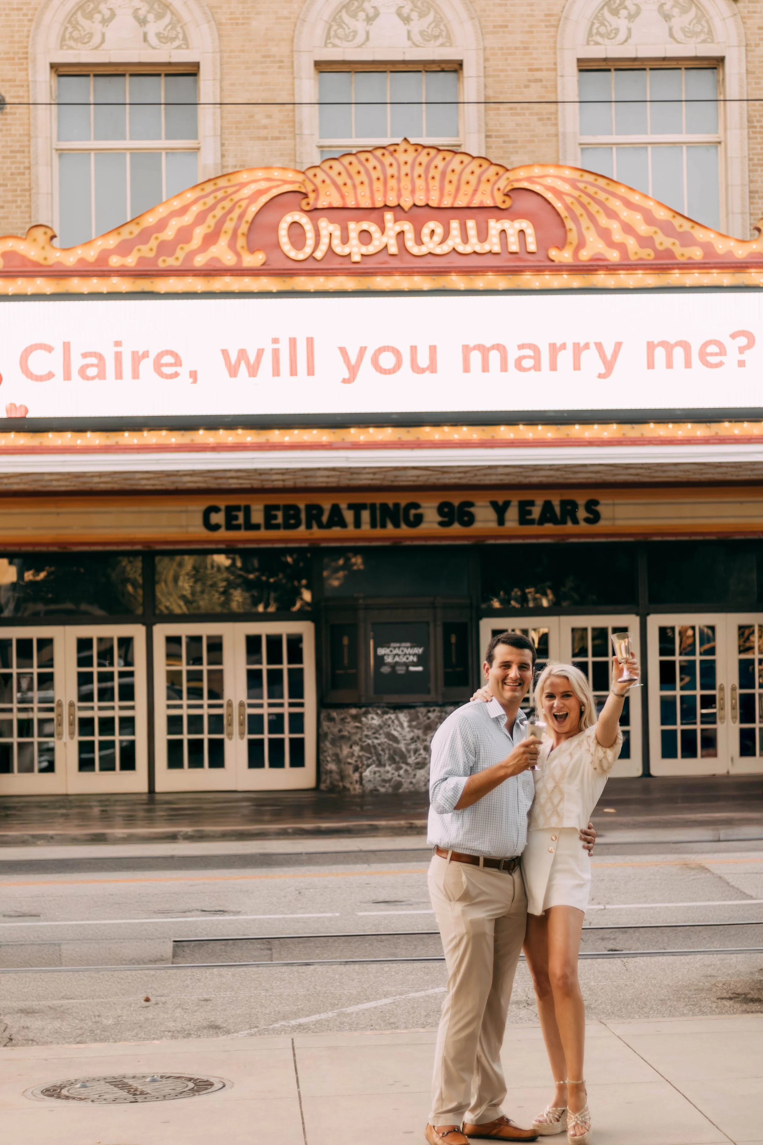 A couple celebrating outside the Orpheum theater, holding drinks and smiling. Behind them, a marquee with a message asking Claire if she will marry the speaker, and a sign indicating they are celebrating 96 years.