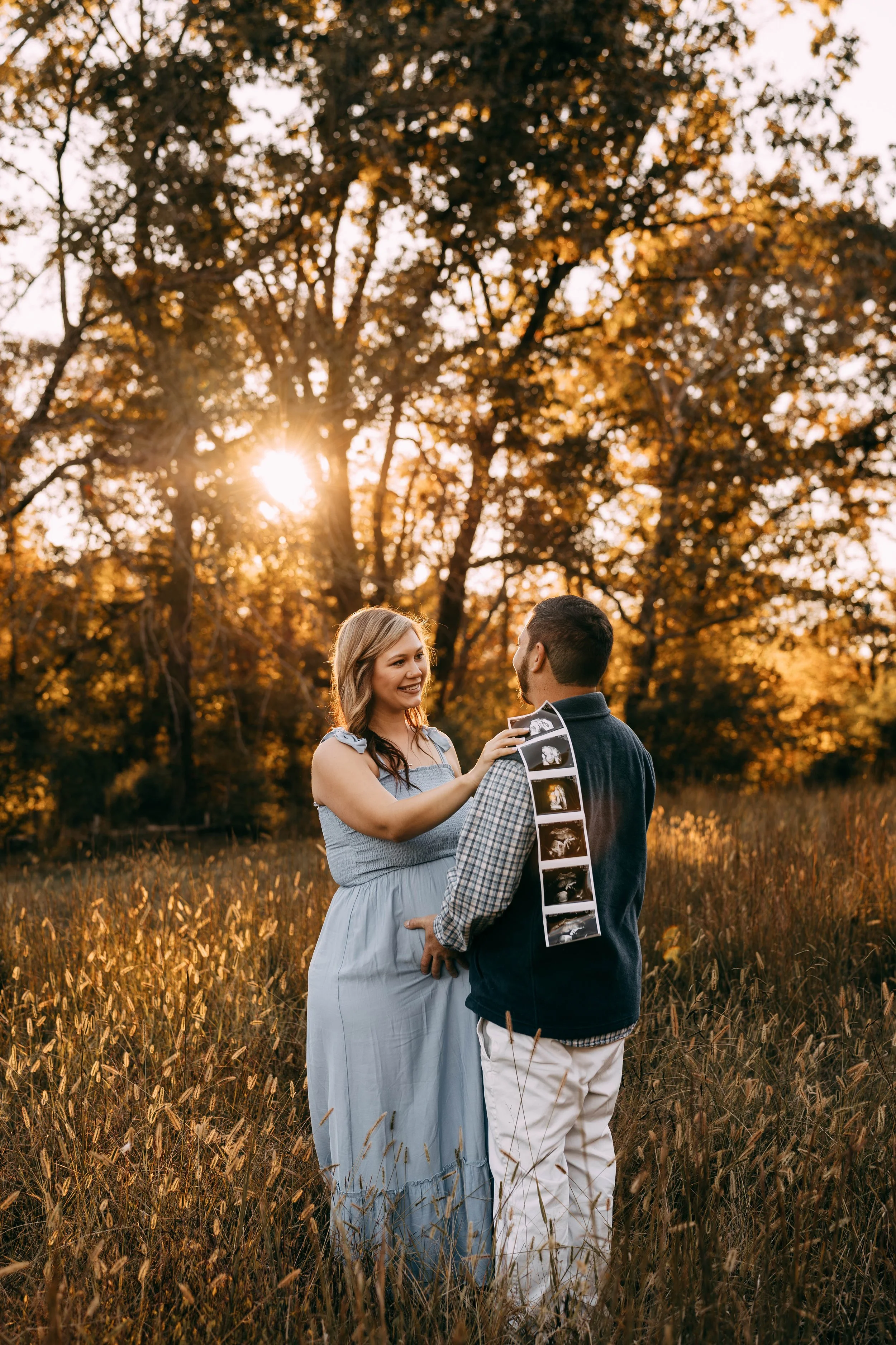 A pregnant woman and a man are standing in a field with tall grass, holding ultrasound images, during sunset with orange filtering through trees in the background.