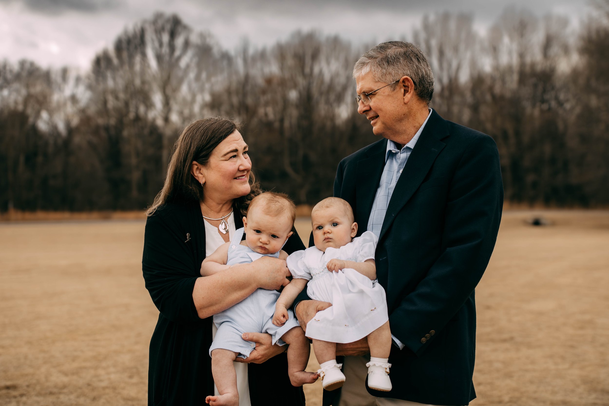 A woman and a man holding two babies outdoors in a park, with trees in the background.