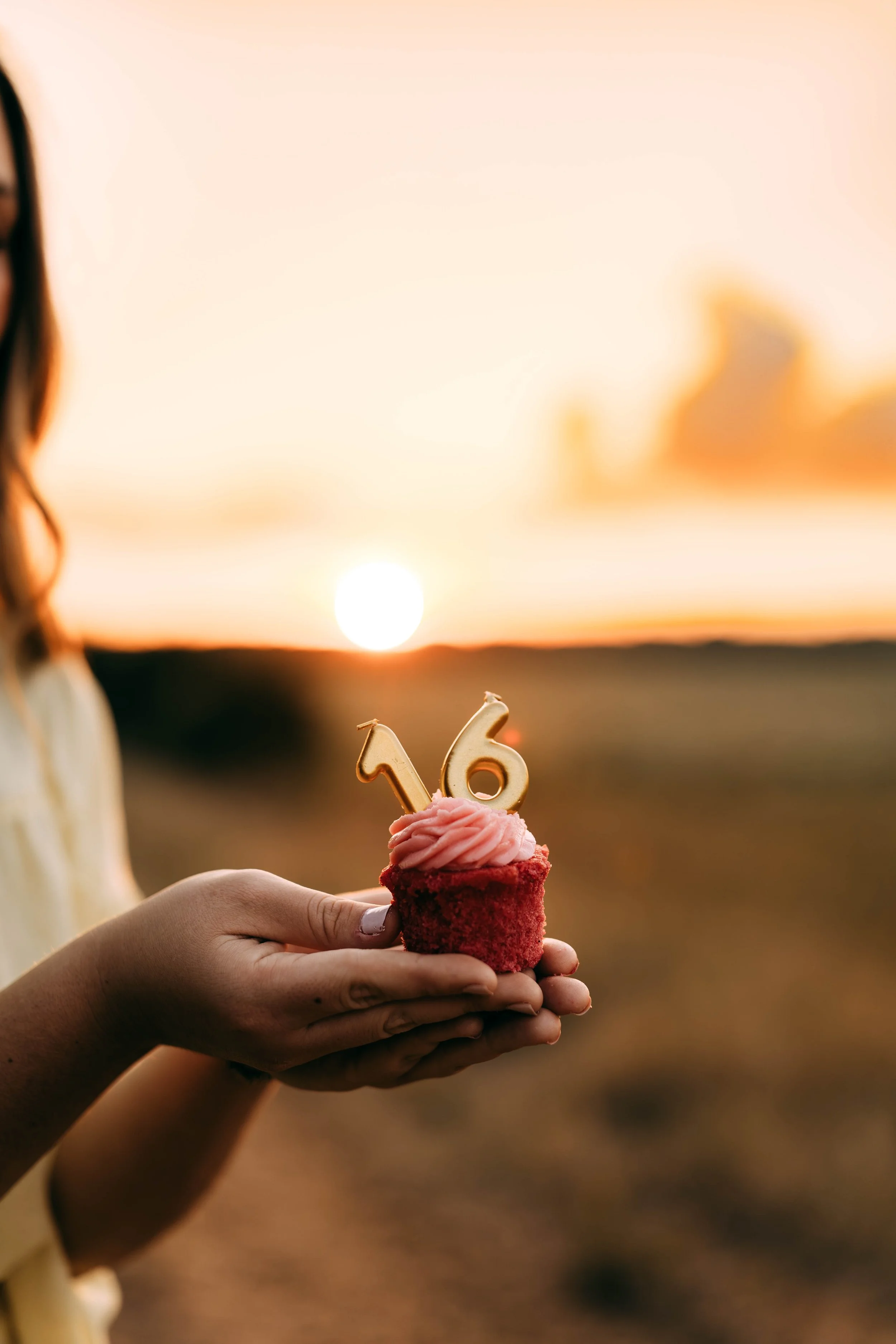 A person holding a cupcake with a pink frosting and a gold candle shaped as the number 16 against a sunset background.