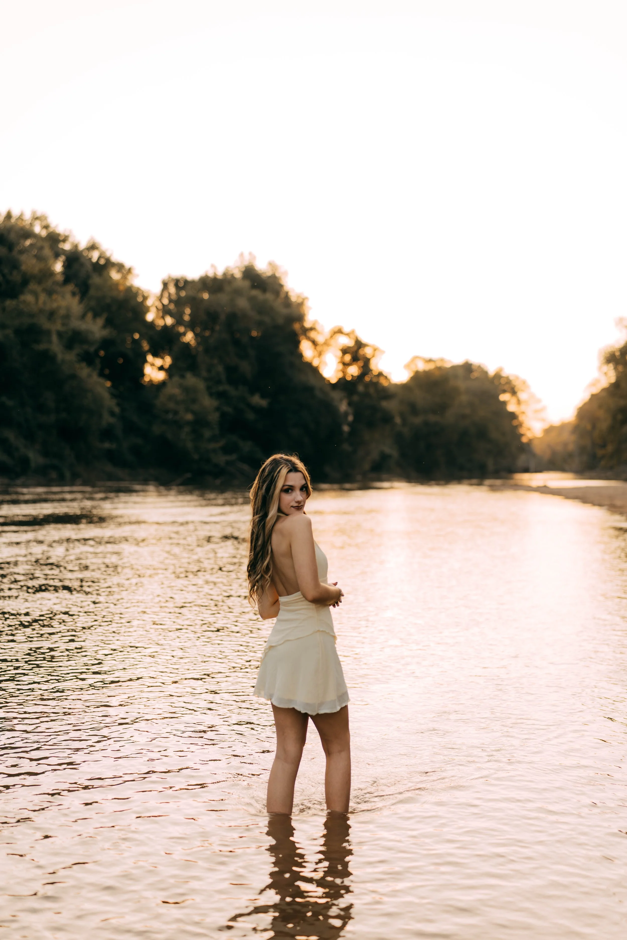 A woman stands in a river at sunset, looking back over her shoulder, wearing a light-colored dress.