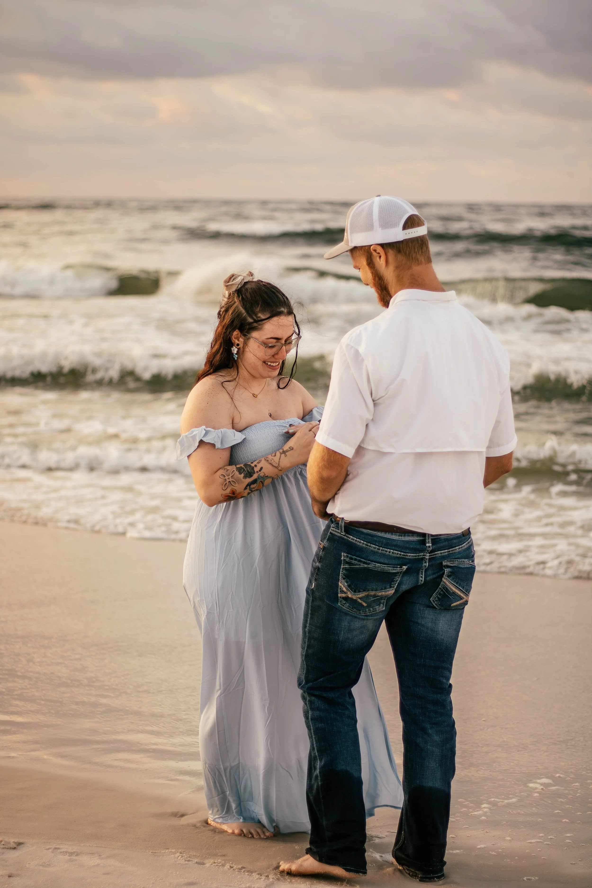 A couple standing on the beach during sunset, with waves in the background, the woman is dressed in a light blue off-shoulder dress with tattoos on her arm, and the man is in a white shirt, jeans, and a white cap, looking at the woman, smiling, durin