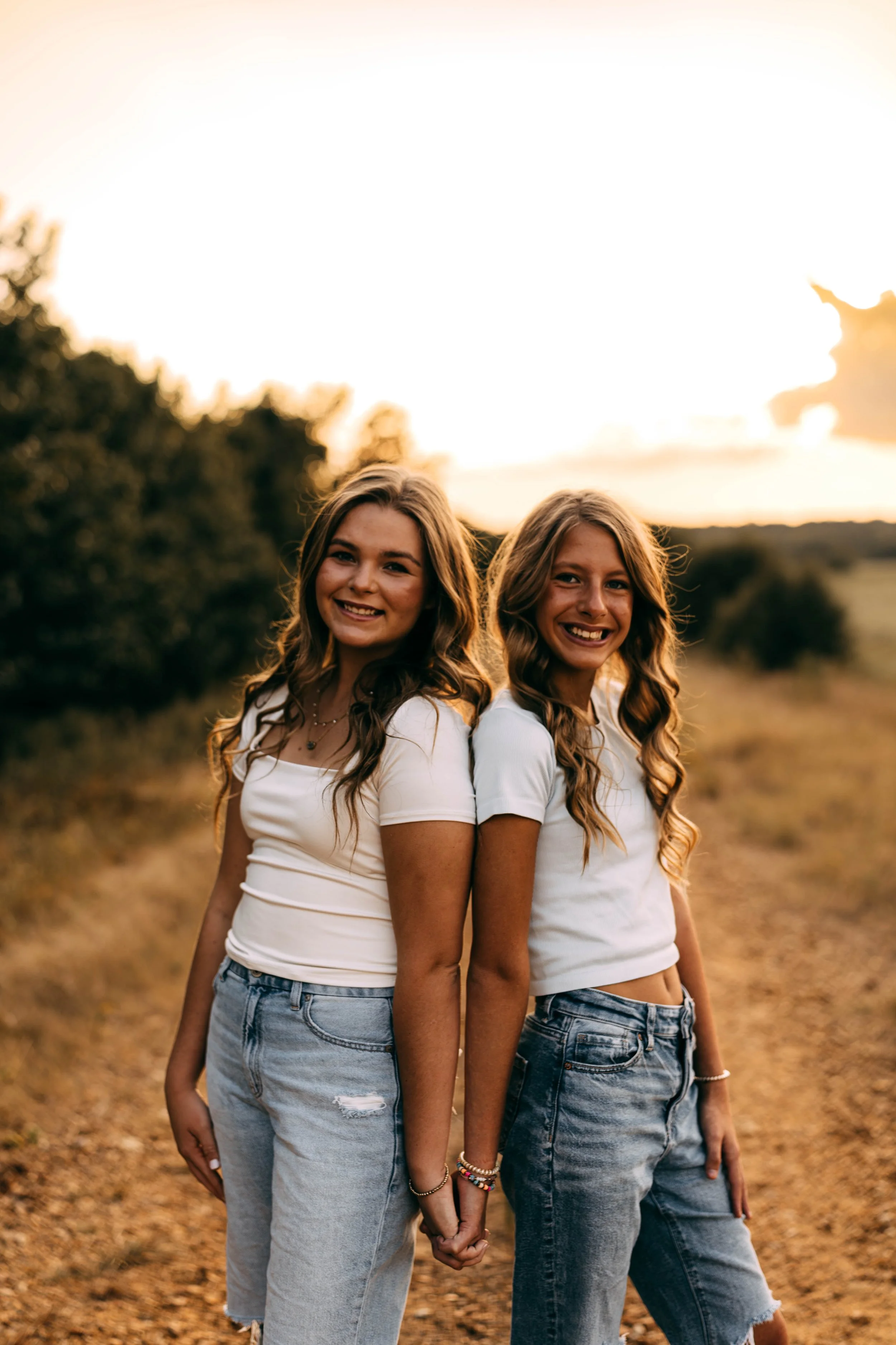 Two young women standing back to back, holding hands, outdoors during sunset, smiling, wearing white t-shirts and jeans, in a natural setting with trees and a dirt path.