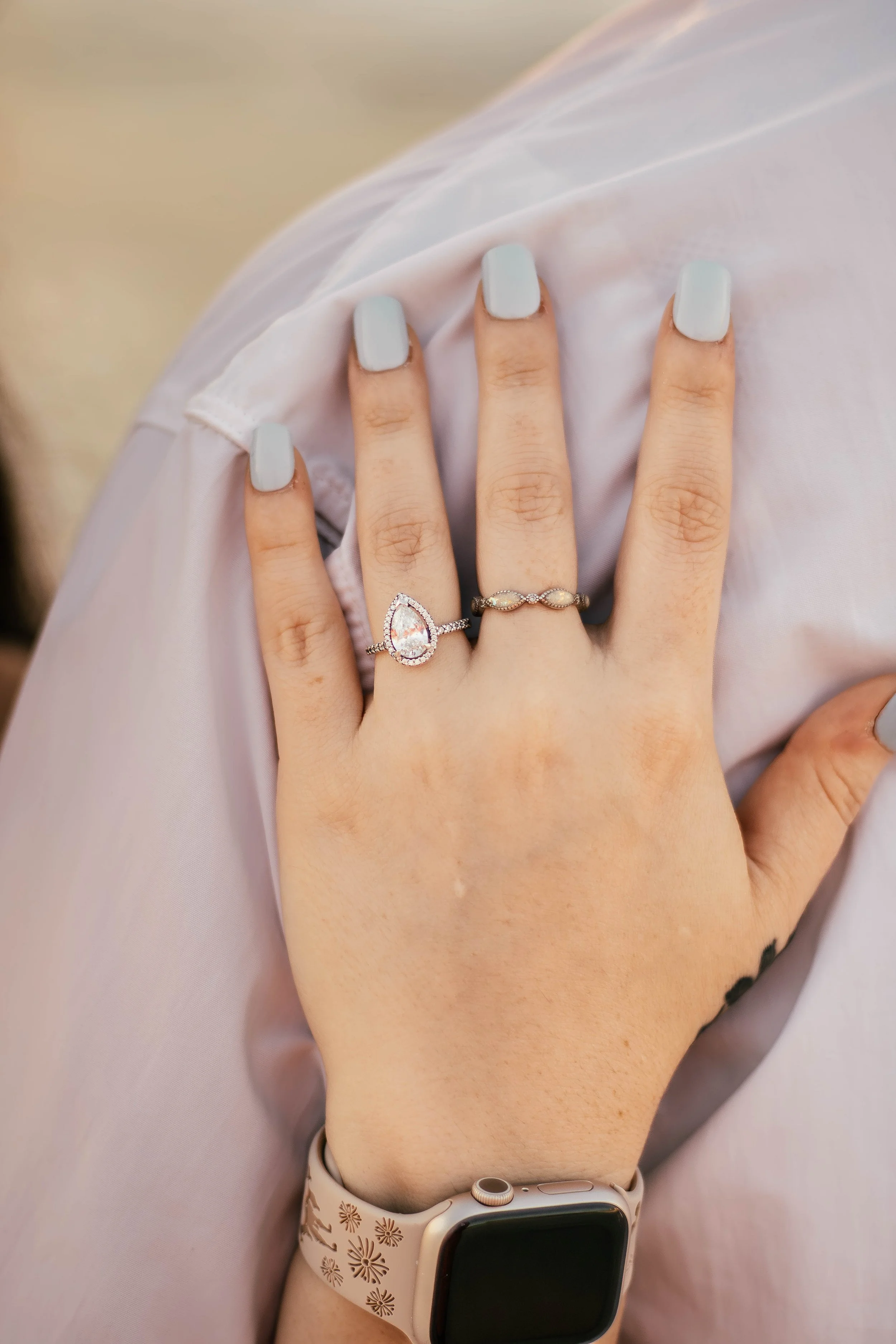 Close-up of a woman's left hand resting on her knee, with two rings on her middle finger, one with a pear-shaped diamond and a silicone smartwatch on her wrist.