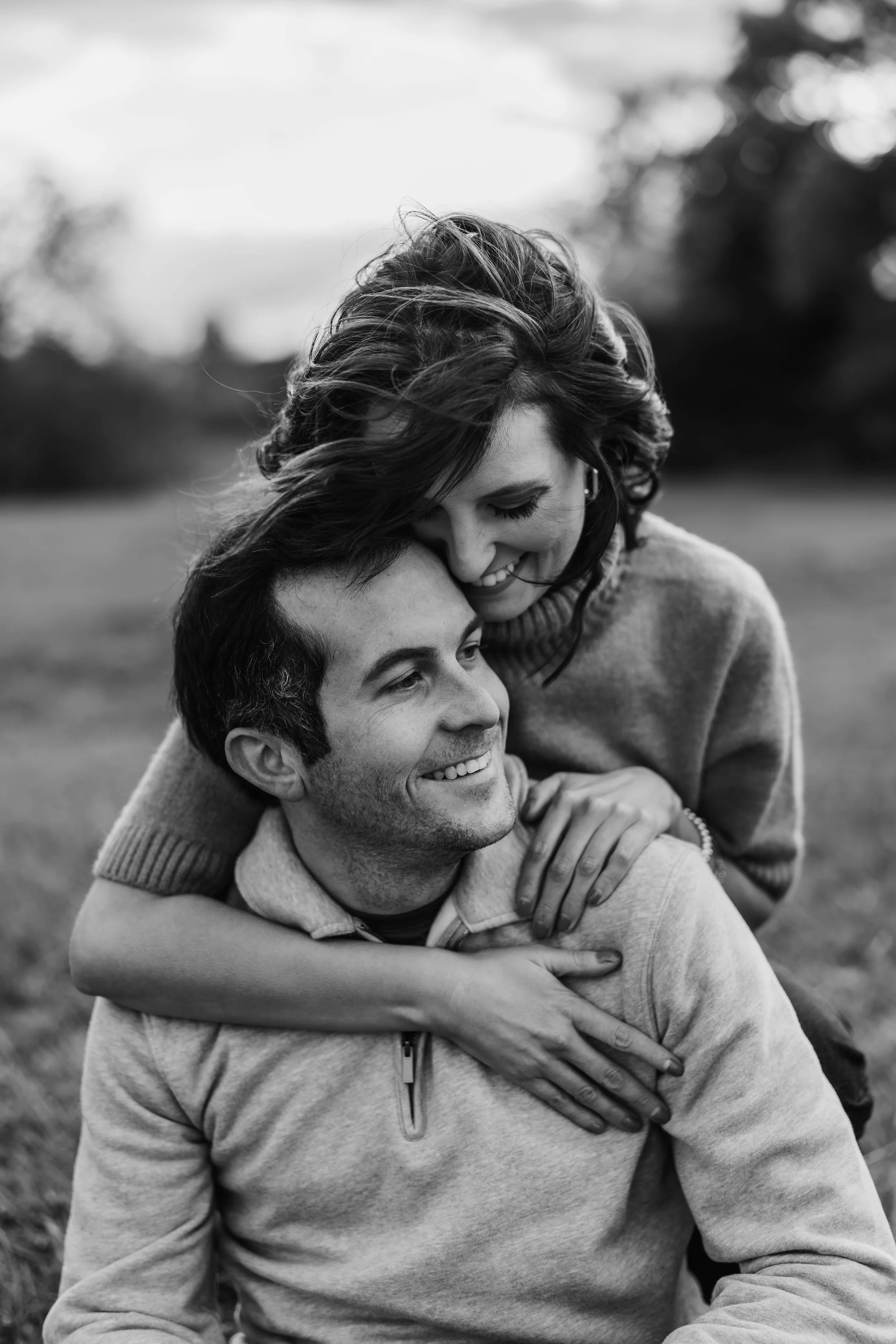 A black and white photo of a smiling couple outdoors, with the woman leaning her head on the man's. They are embracing and appear happy.