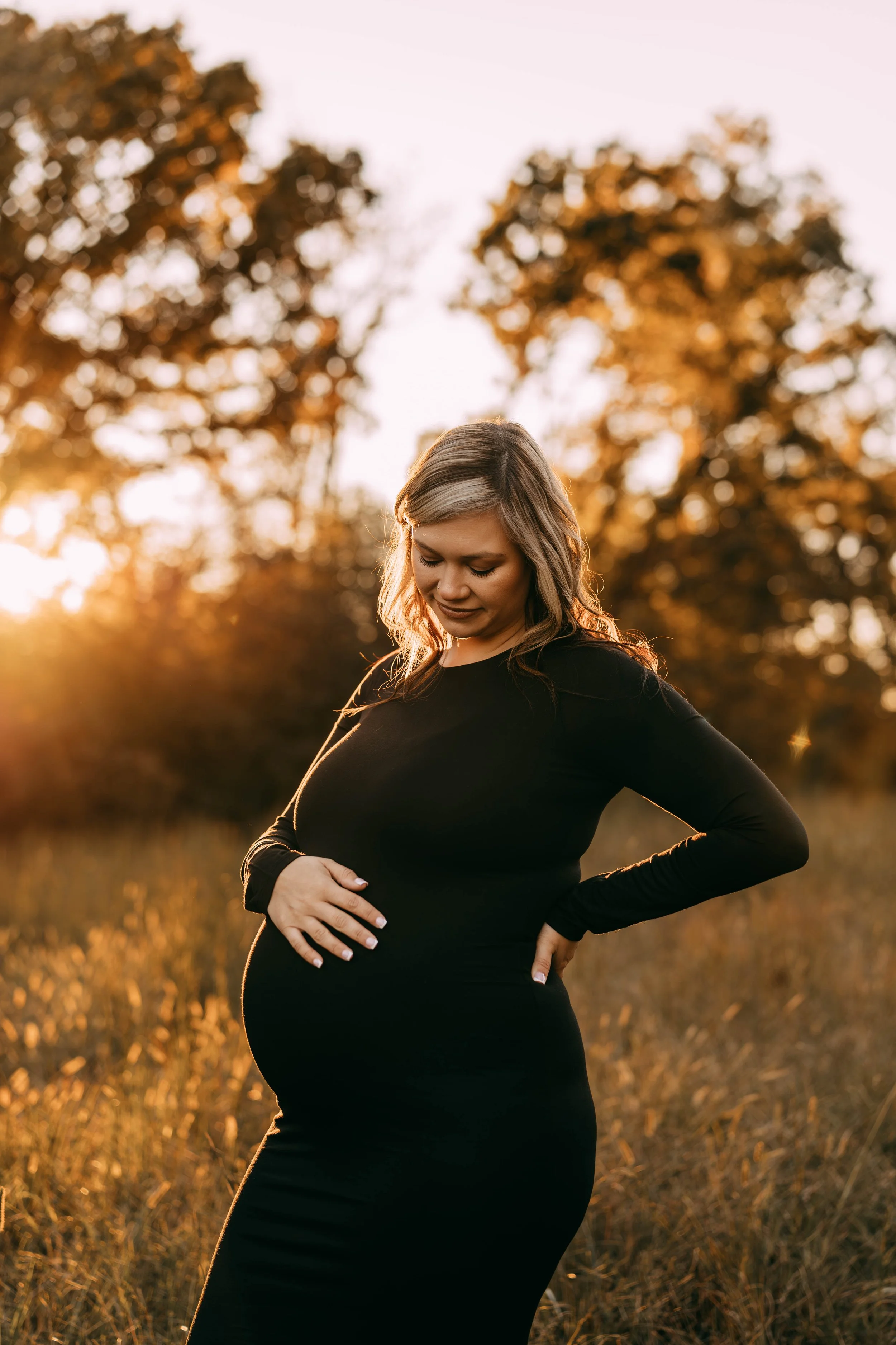 Prettily dressed pregnant woman standing outdoors in a field during sunset, cradling her belly with her hands, with warm golden light and trees in the background.