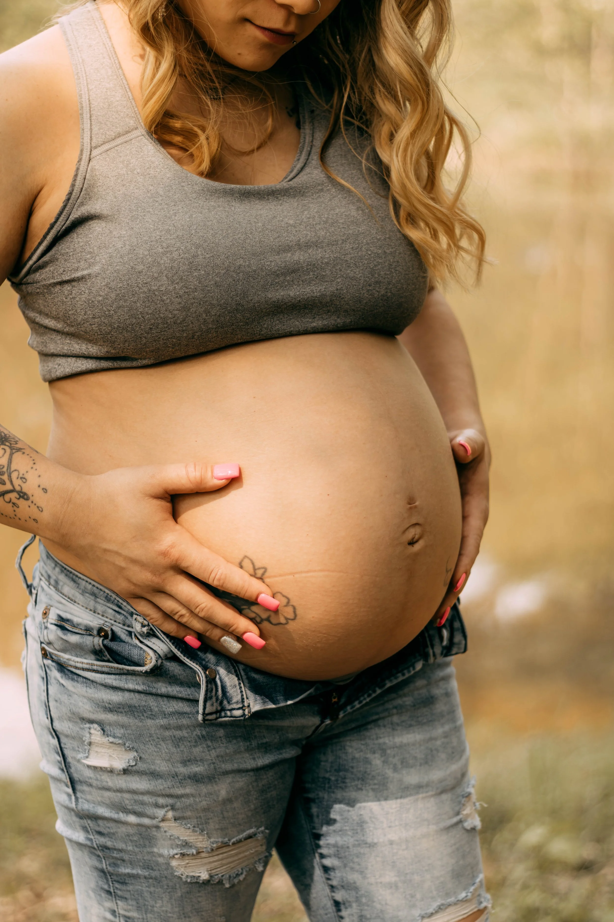 A pregnant woman with blonde hair, wearing a gray tank top and ripped jeans, gently cradling her belly outdoors in a natural setting.