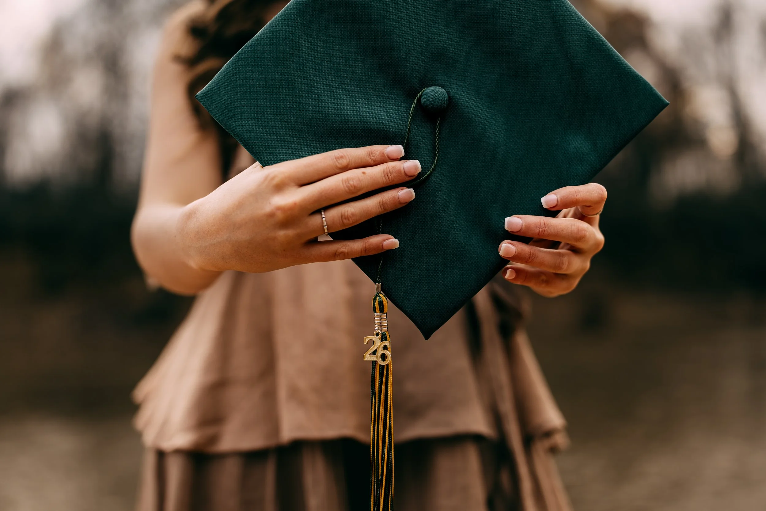 Person holding a green graduation cap with a tassel and a gold '26' ornament, outdoors in natural light.
