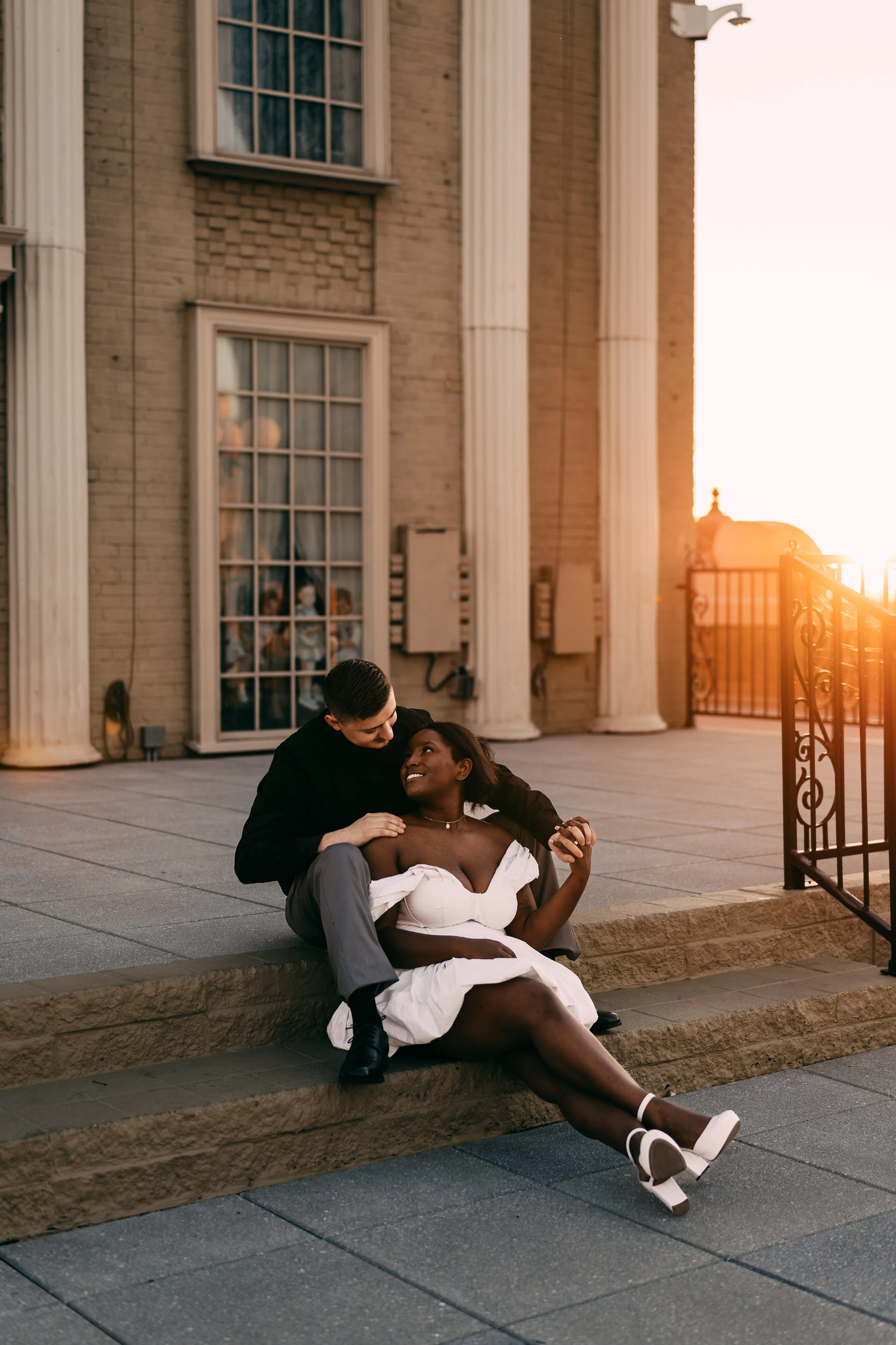 A couple sitting on a step outside a building with large columns at sunset, smiling and looking at each other.