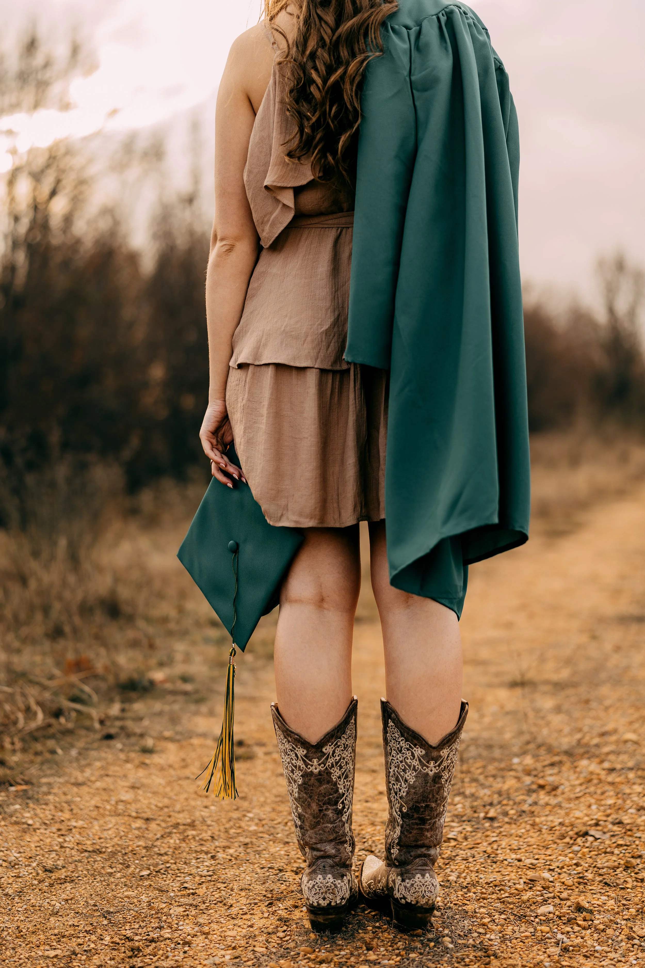 A woman in a brown dress holding a graduation cap in her right hand, standing outdoors on a dirt path with trees in the background.