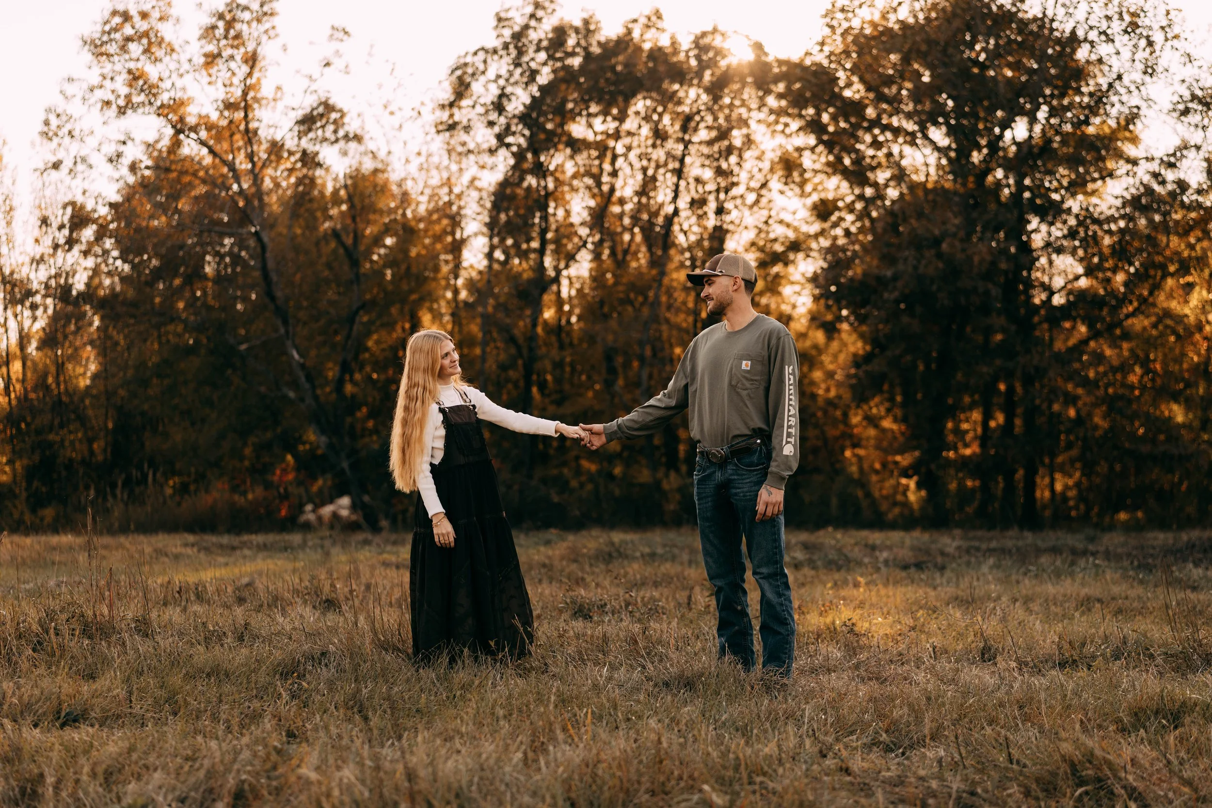A man and a girl standing in a field holding hands at sunset, with autumn trees in the background.
