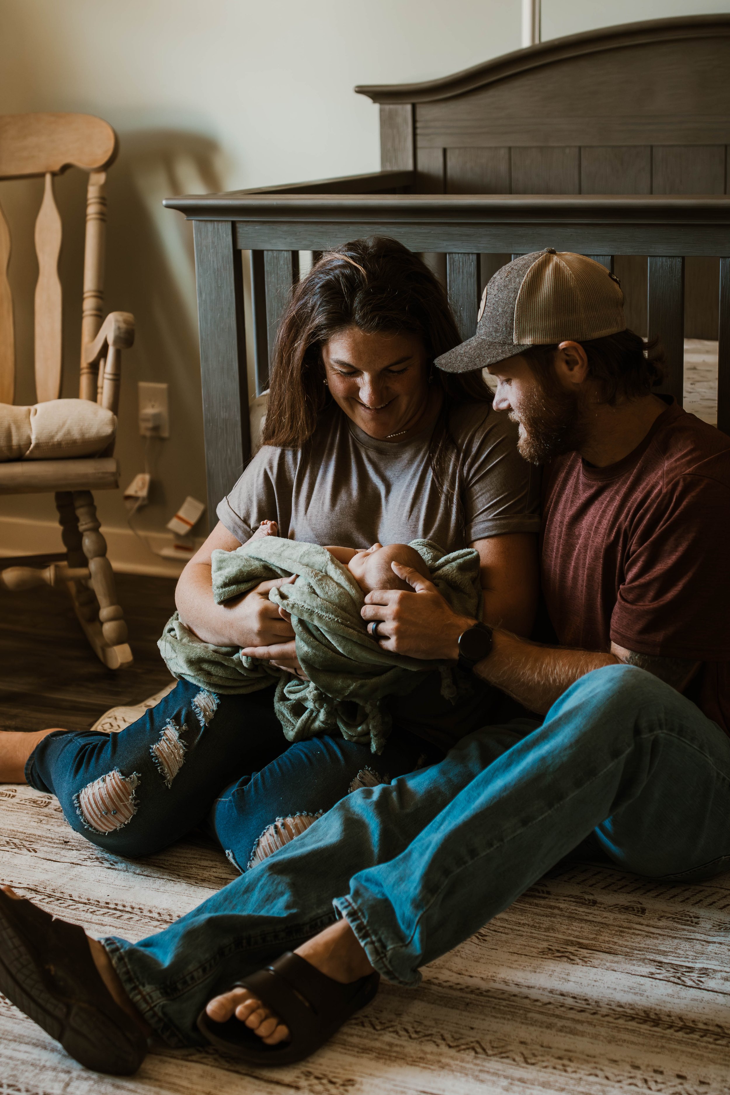 A woman and a man sitting on the floor, holding a newborn baby wrapped in a blanket, in a cozy room with wooden furniture.