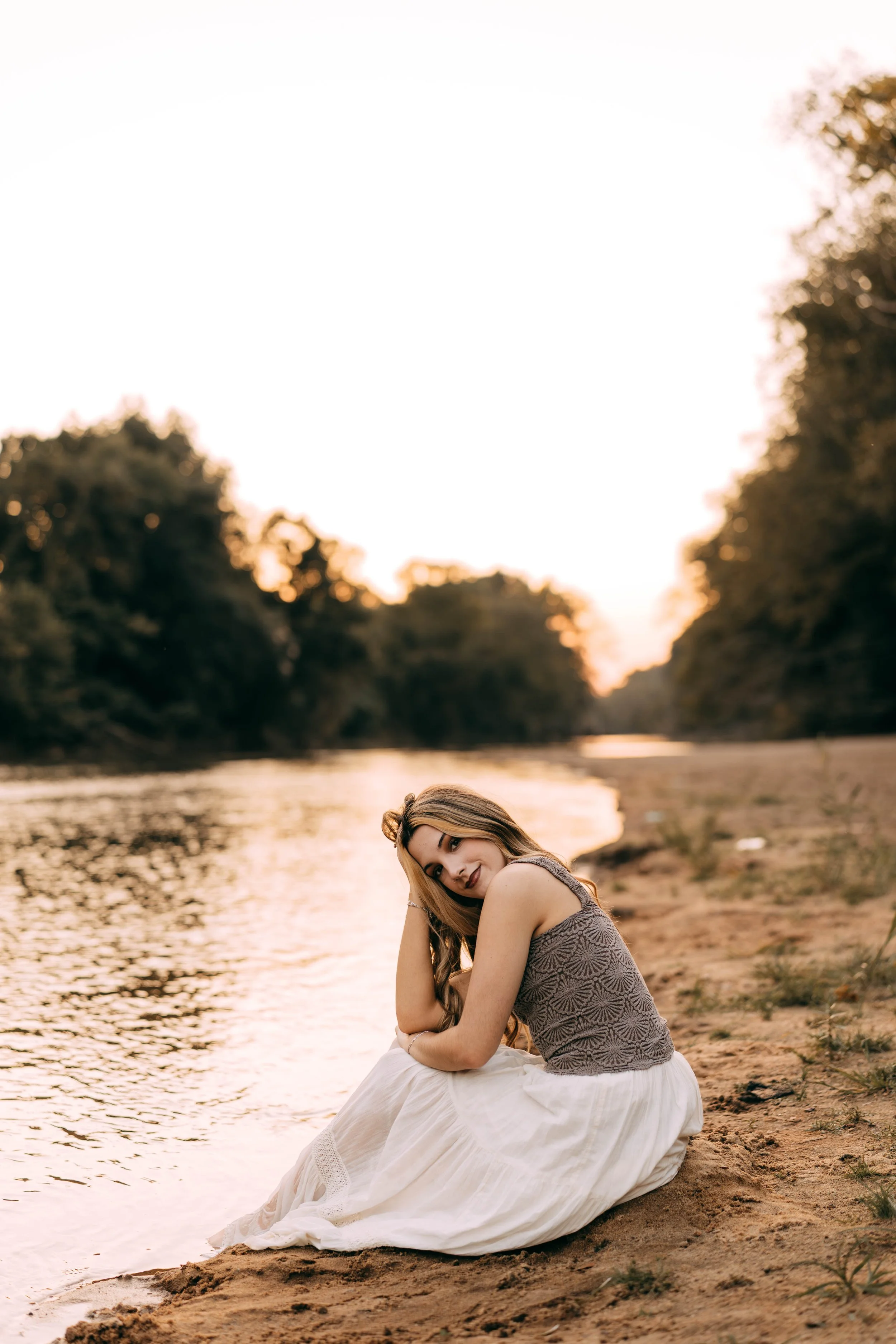 A young woman sitting on a sandy riverbank during sunset, with trees in the background, wearing a gray sleeveless top and a long white skirt, looking at the camera.