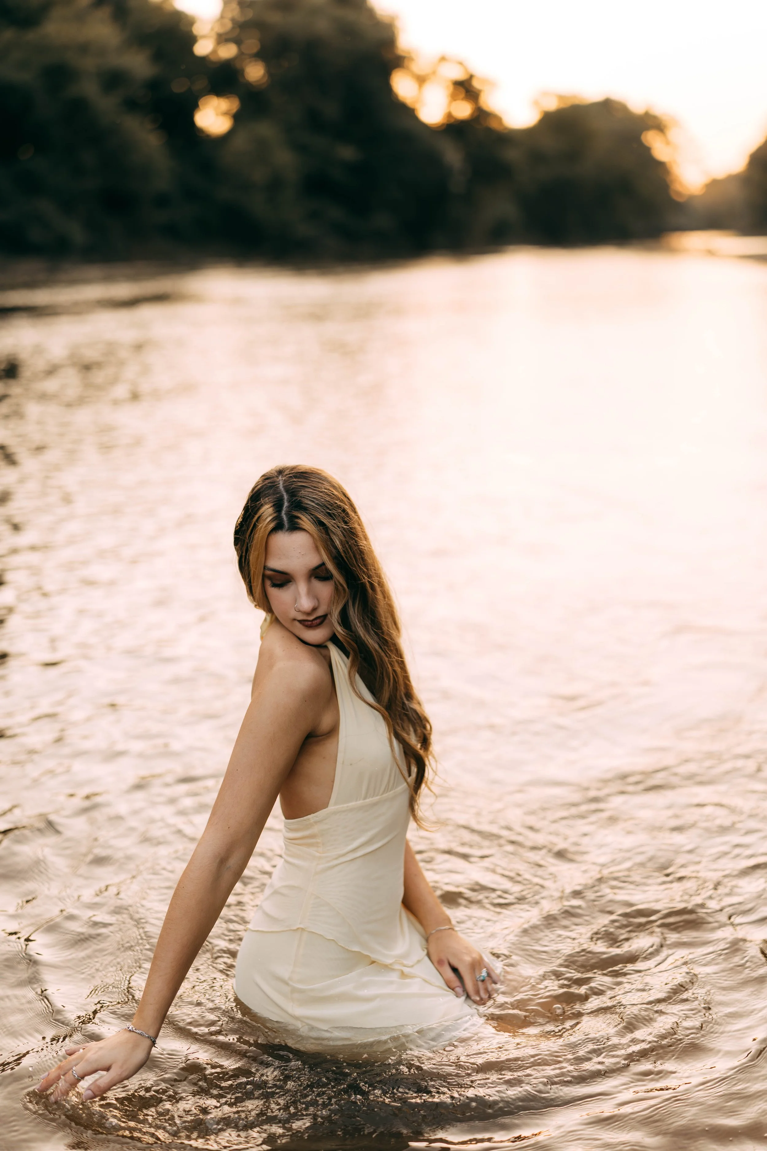 A woman in a cream-colored dress sitting in a river at sunset with her eyes closed and head tilted downward.