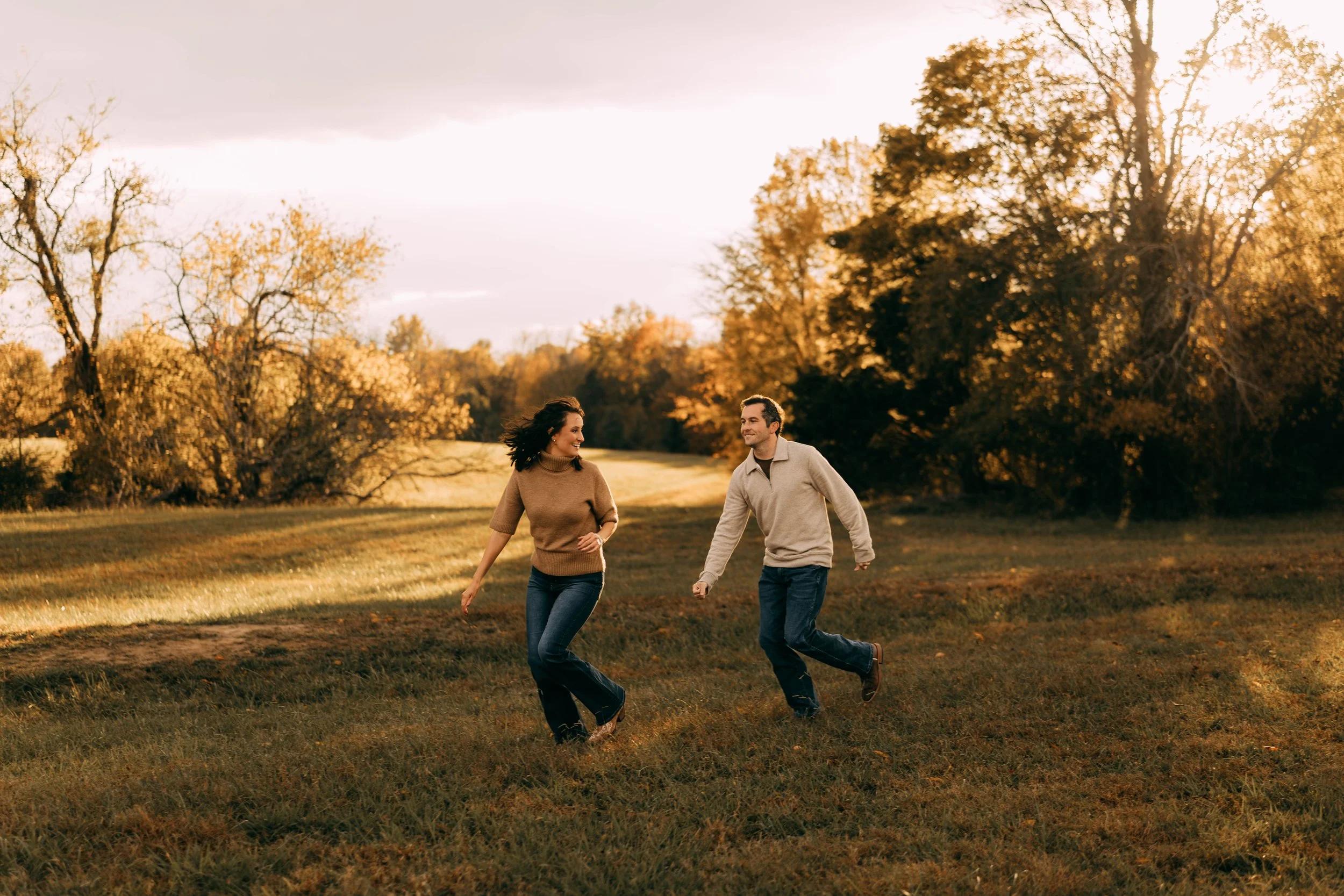 A couple running and smiling in a grassy field during autumn with trees in the background and warm sunlight.