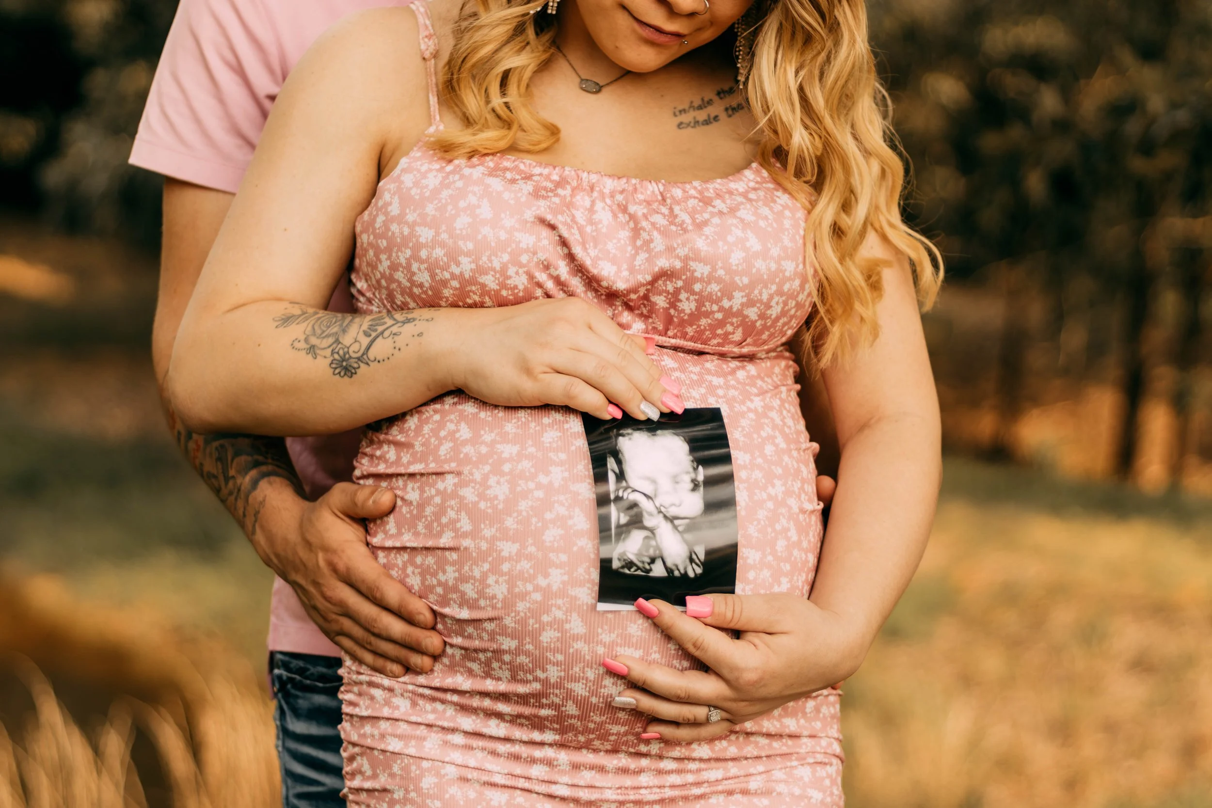 Pregnant woman holding an ultrasound photo on her belly, with her partner's hands around her waist, outdoors in a forest setting.