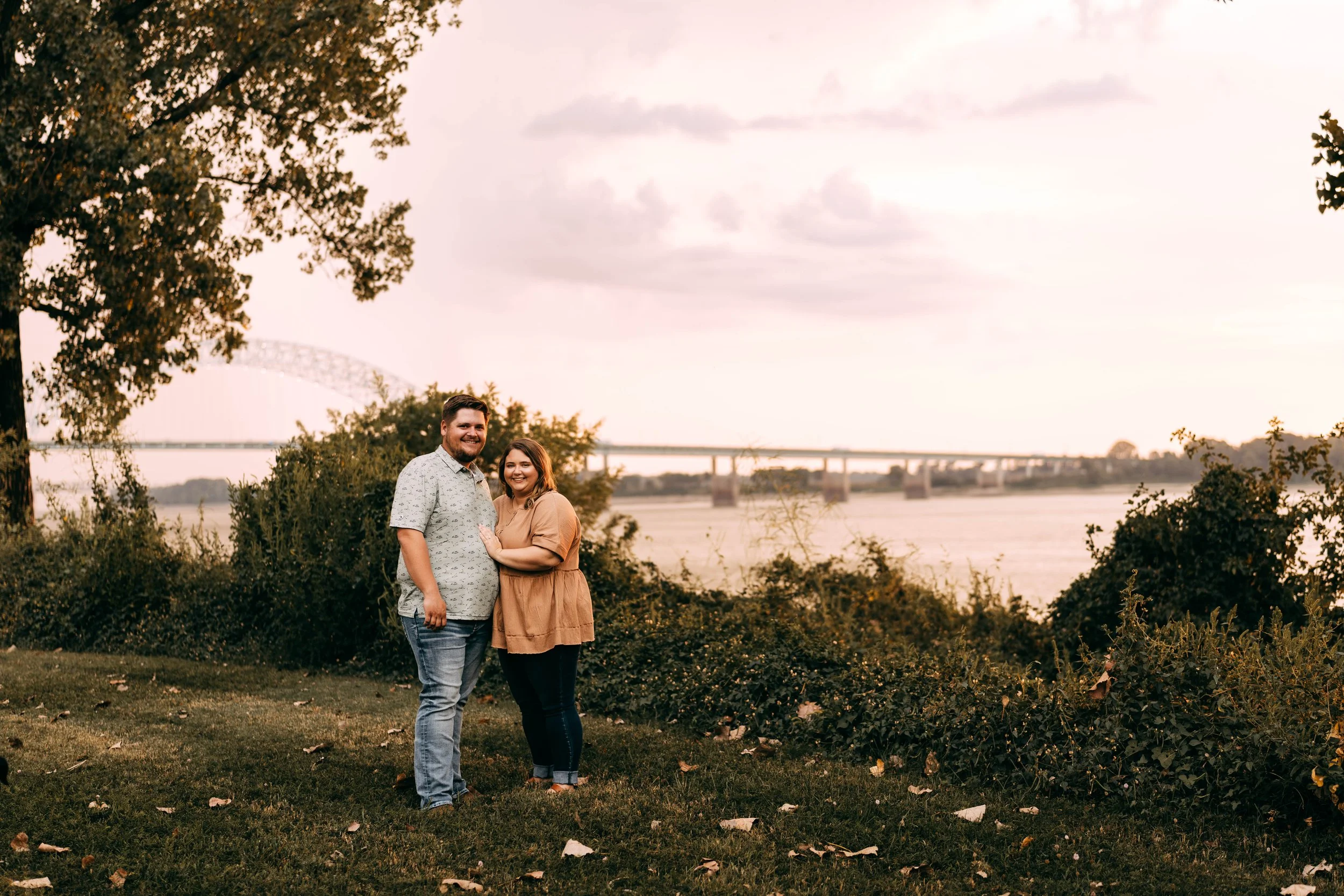 A couple standing outdoors near a river during sunset, smiling at the camera, with trees and a bridge in the background.
