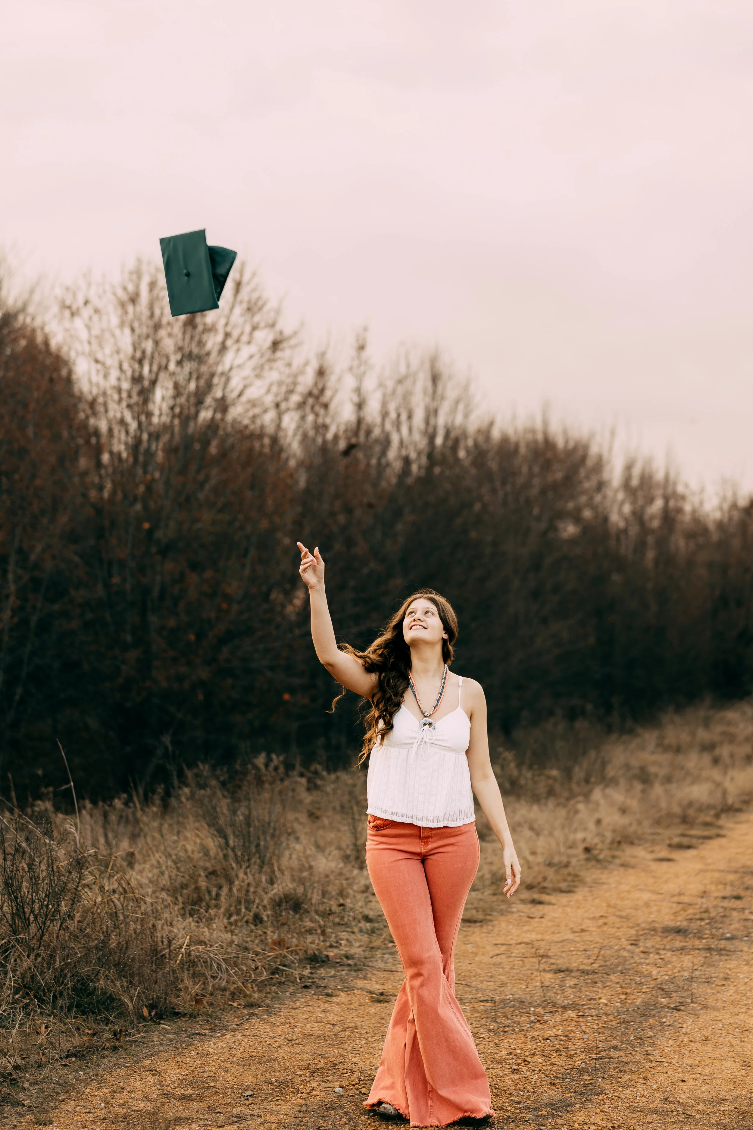 A young woman in a white sleeveless top and orange bell-bottom jeans is walking on a dirt path, throwing her graduation cap into the air, with a background of trees and pink sky.
