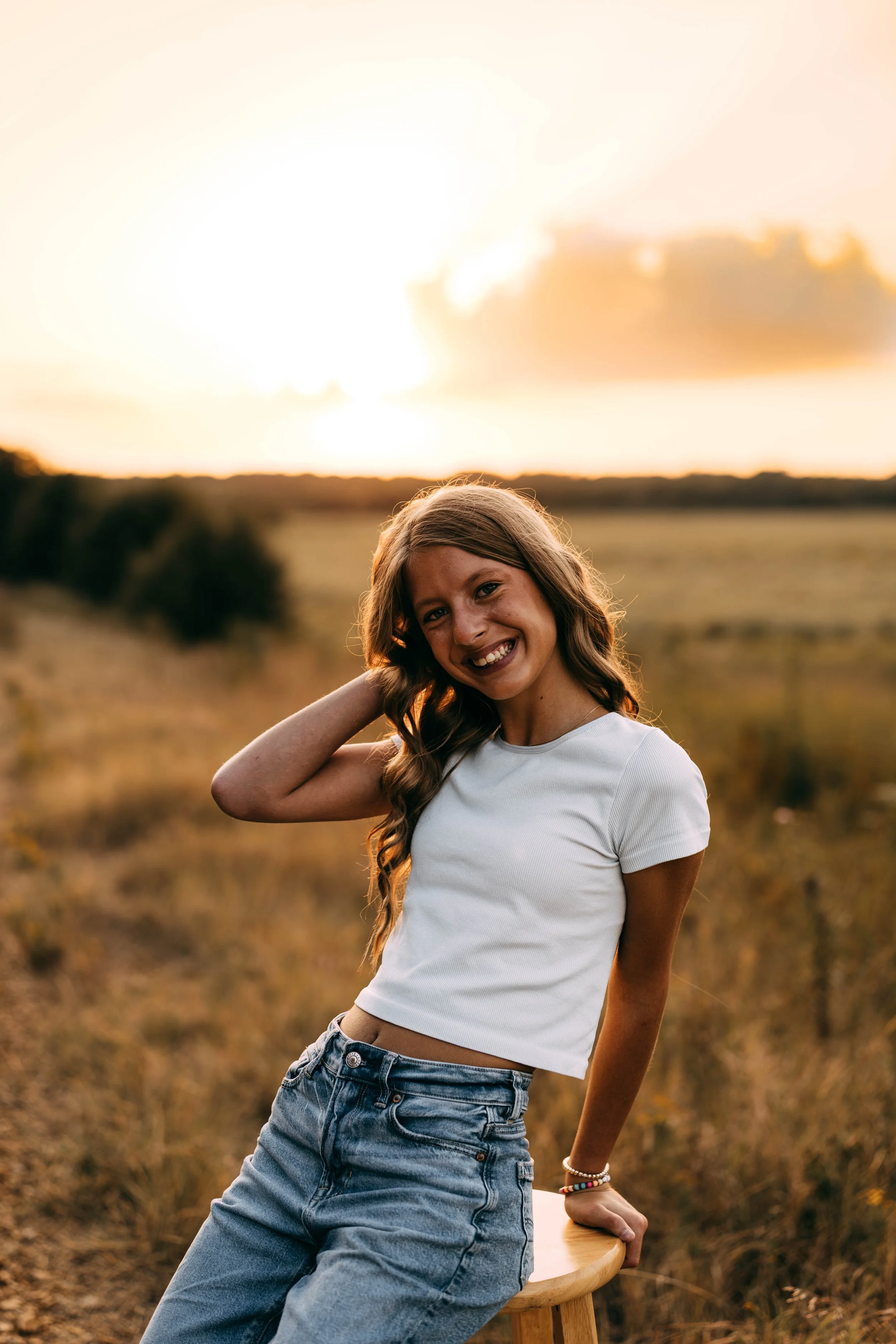 A young girl with long, wavy hair smiling outdoors during sunset, wearing a white crop top and jeans, standing in a field.