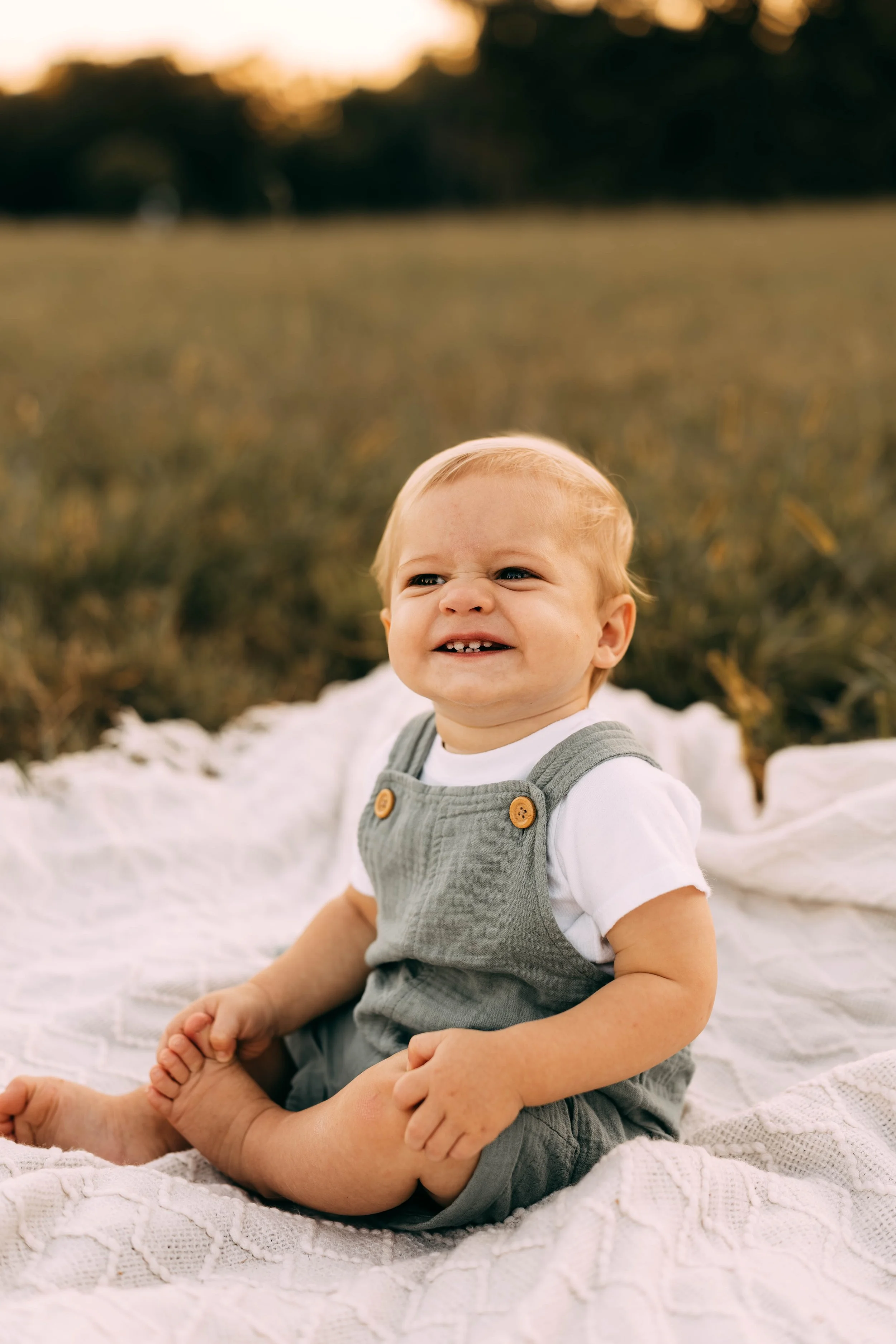 A young child with blonde hair and a big smile sitting on a white blanket outdoors during sunset