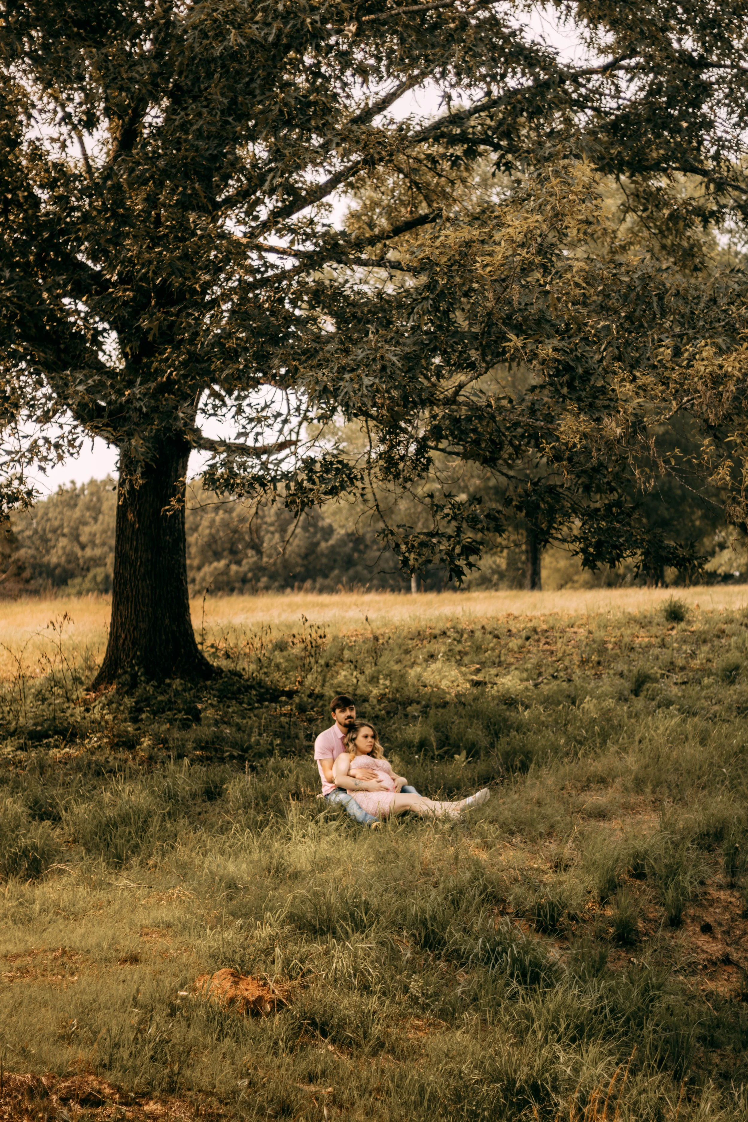A couple, a man and a woman, sitting on the grass under a large tree, with the woman resting on the man's lap, in a grassy park setting.