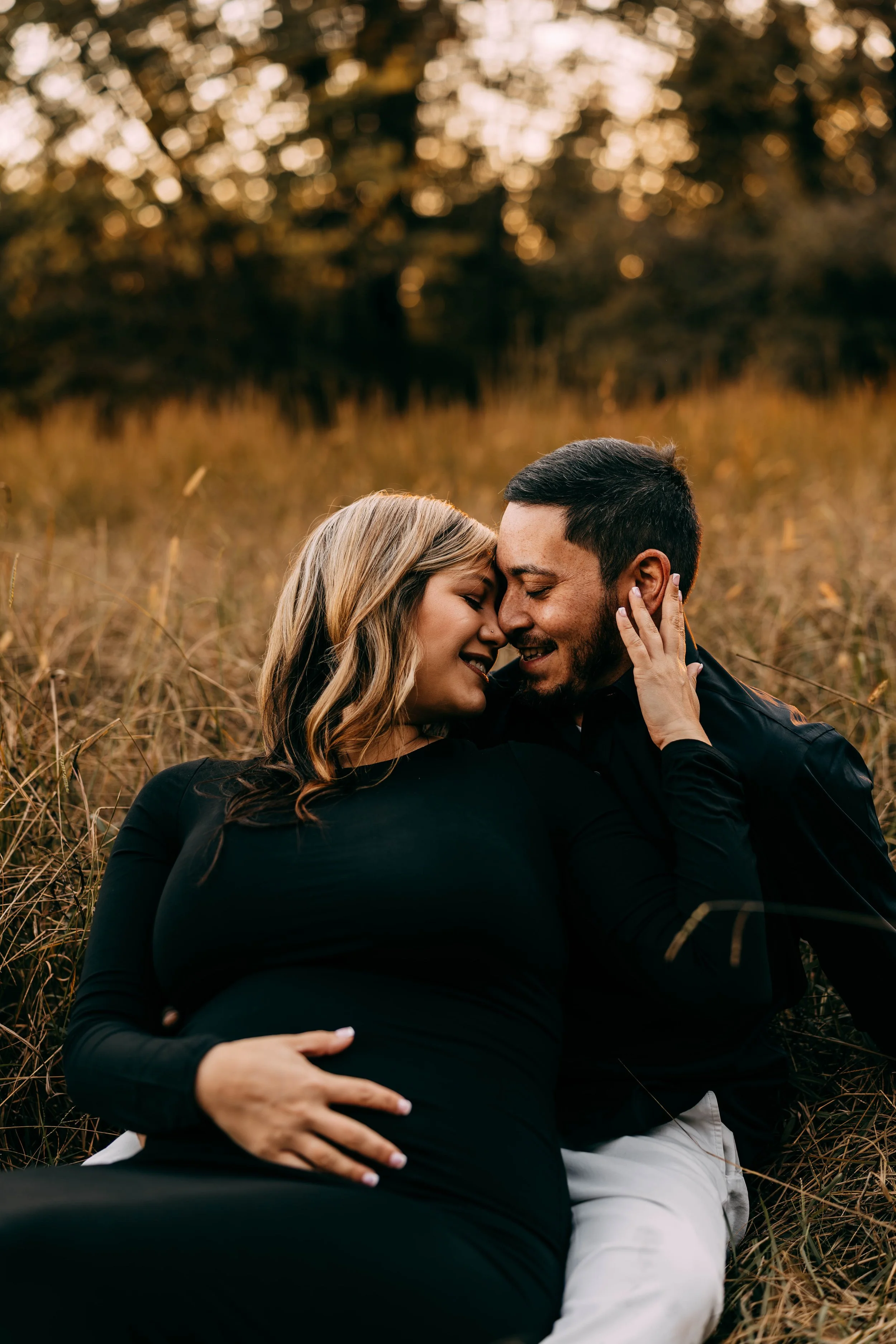 A couple sits outdoors in a field at sunset, sharing a close, intimate moment with their foreheads touching.