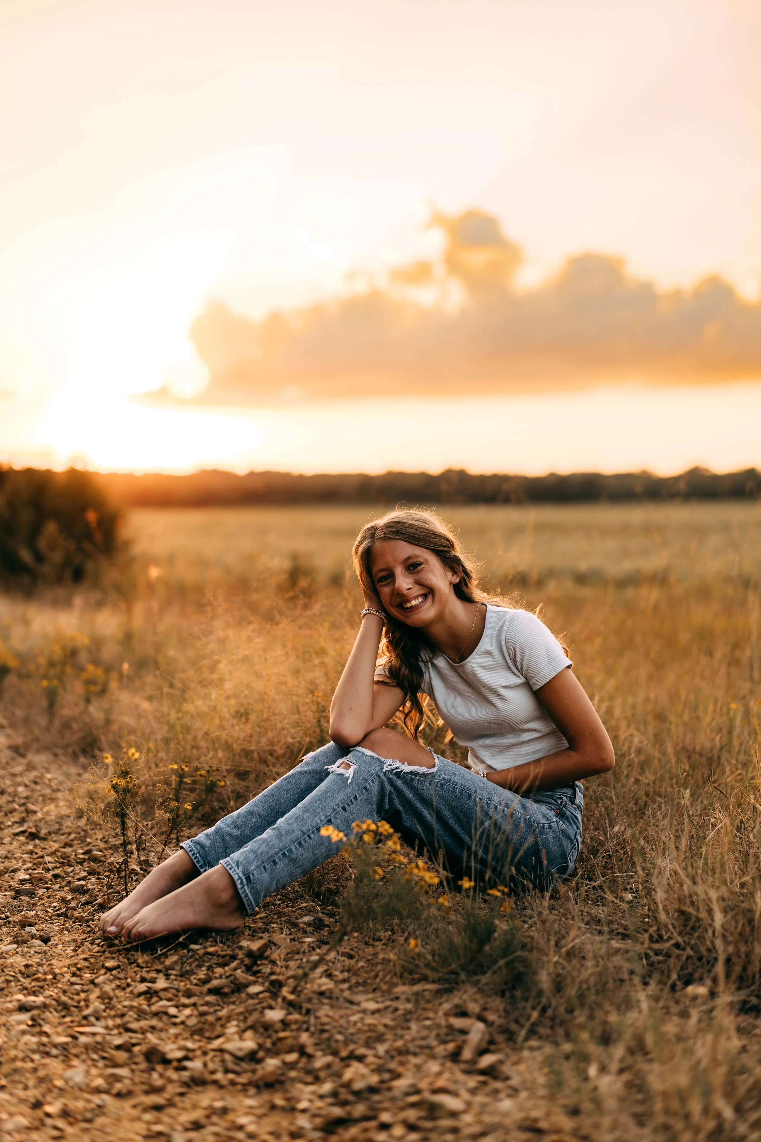 A young woman sitting barefoot on a dirt path in a field during sunset, smiling and wearing a white t-shirt and ripped jeans.