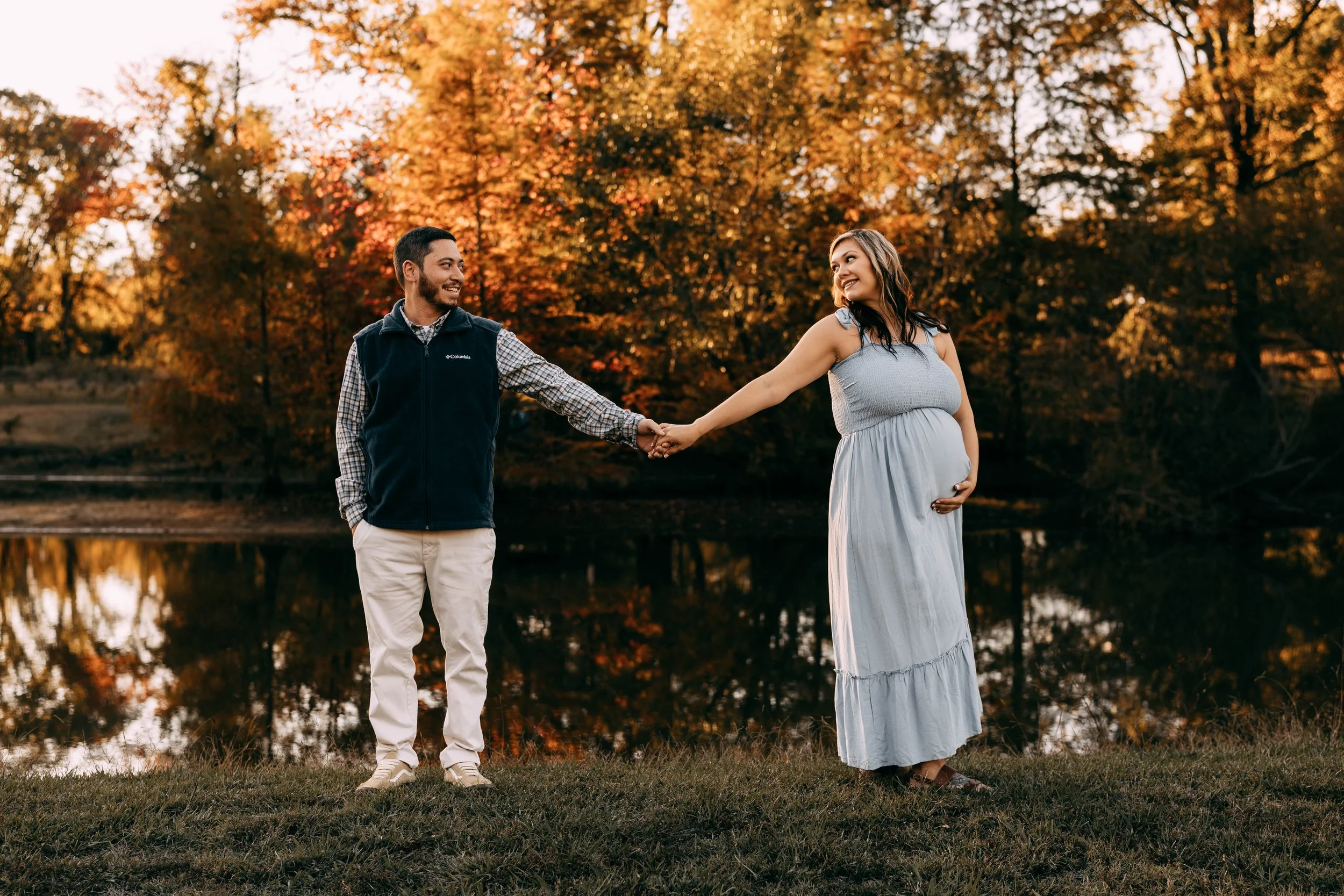 A pregnant woman in a light blue dress holding her belly stands by a lake at sunset, holding hands with a man in a navy vest and khaki pants, both smiling, with autumn trees in the background.