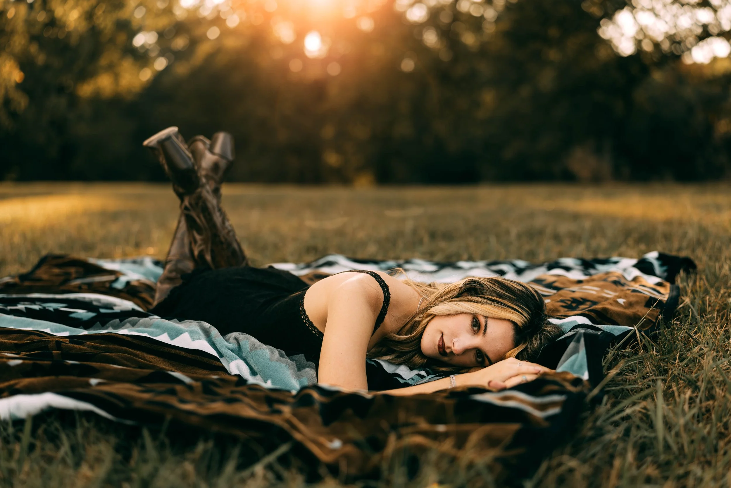 Woman lying on her stomach on a blanket in a grassy field at sunset, looking at the camera.