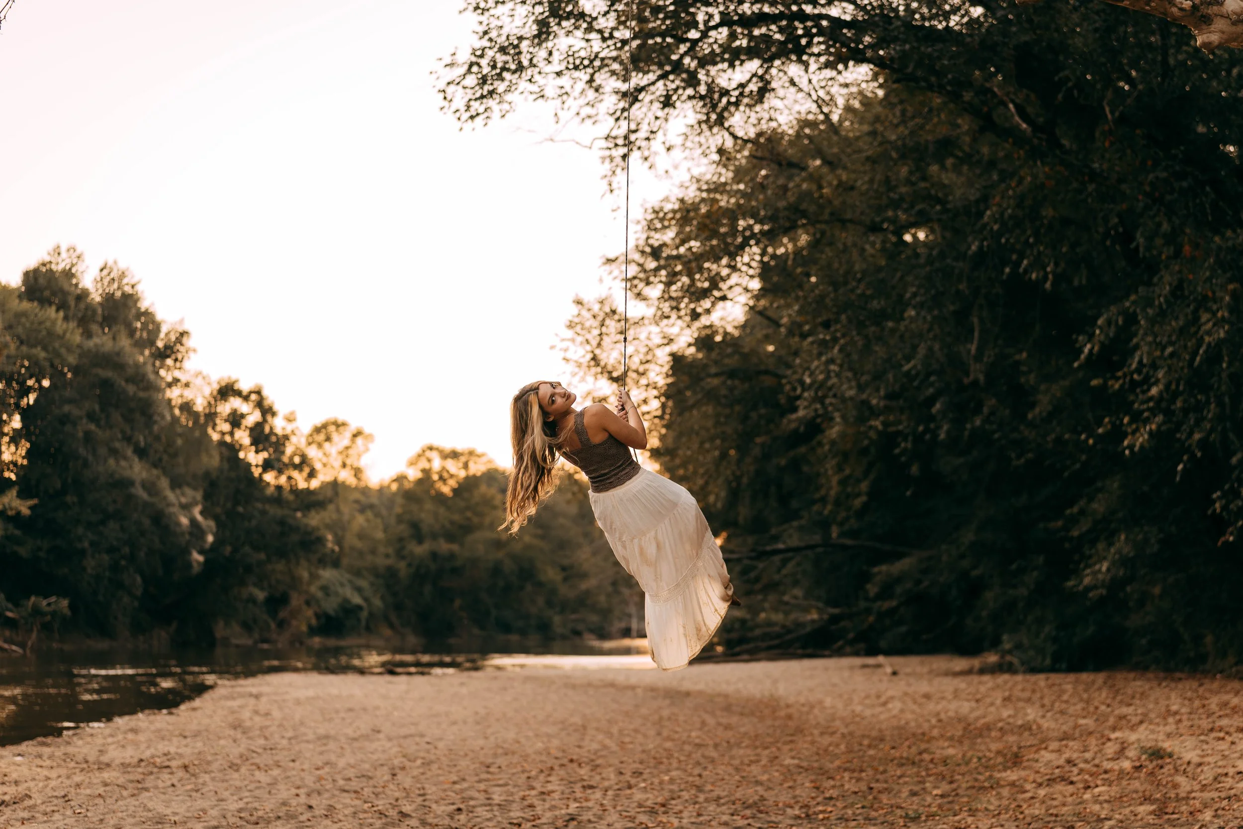 A woman on a swing over a river at sunset surrounded by trees.