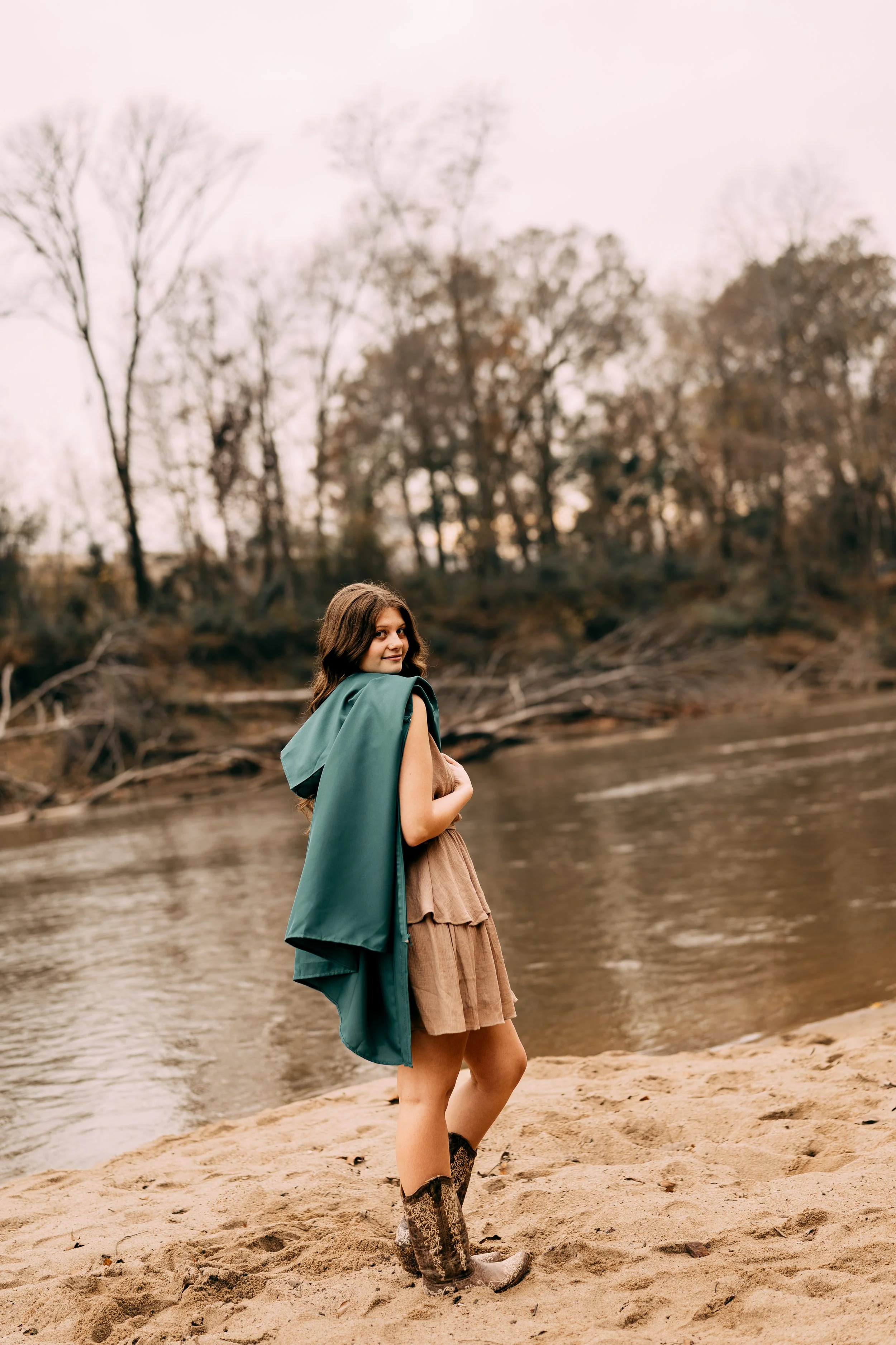 Young woman standing on sandy riverbank in fall, wearing a brown dress, patterned boots, and holding a teal jacket over her shoulder, with leafless trees in the background.