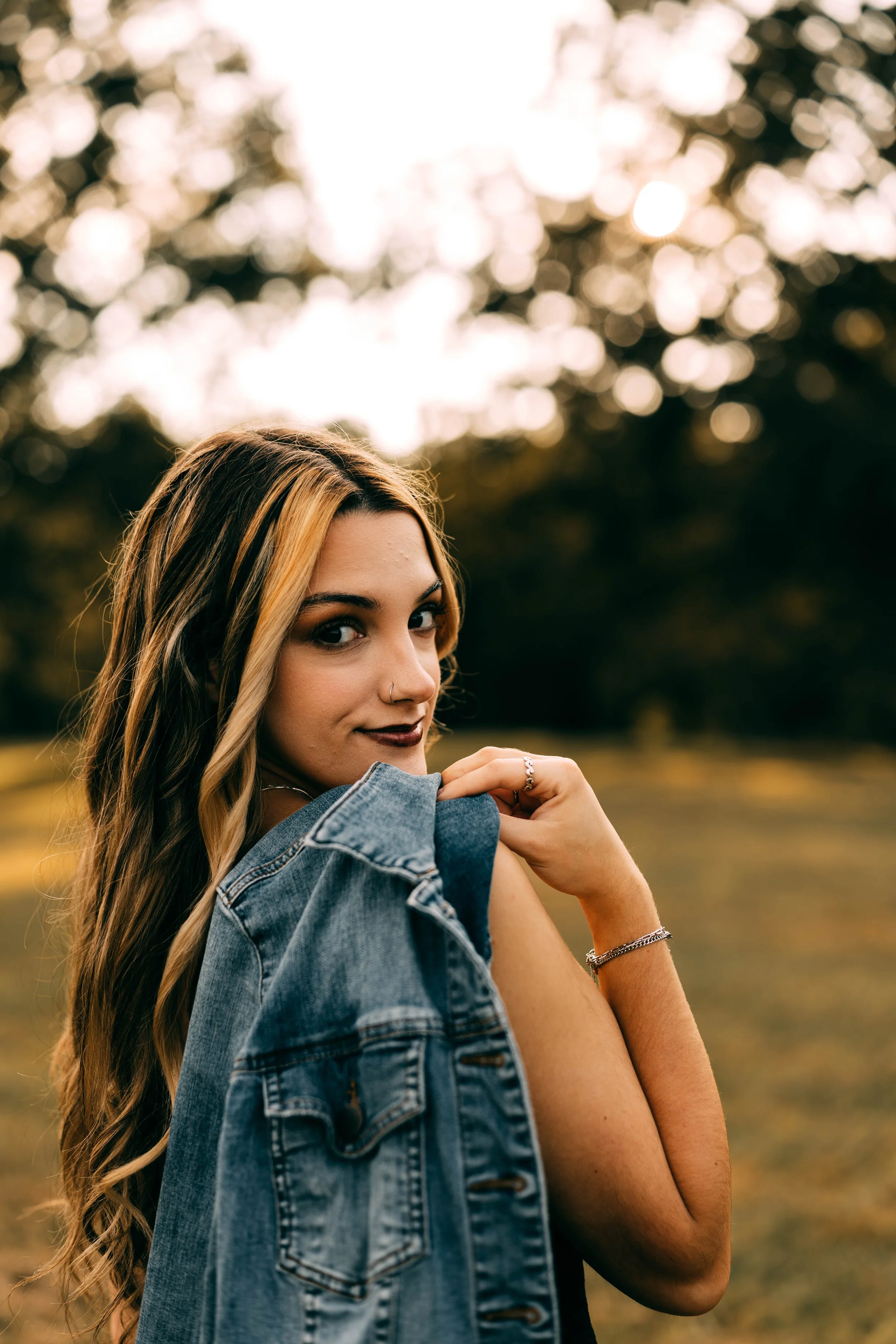 Young woman with long wavy hair holding a denim jacket over her shoulder outdoors in a park with trees in the background during sunset.