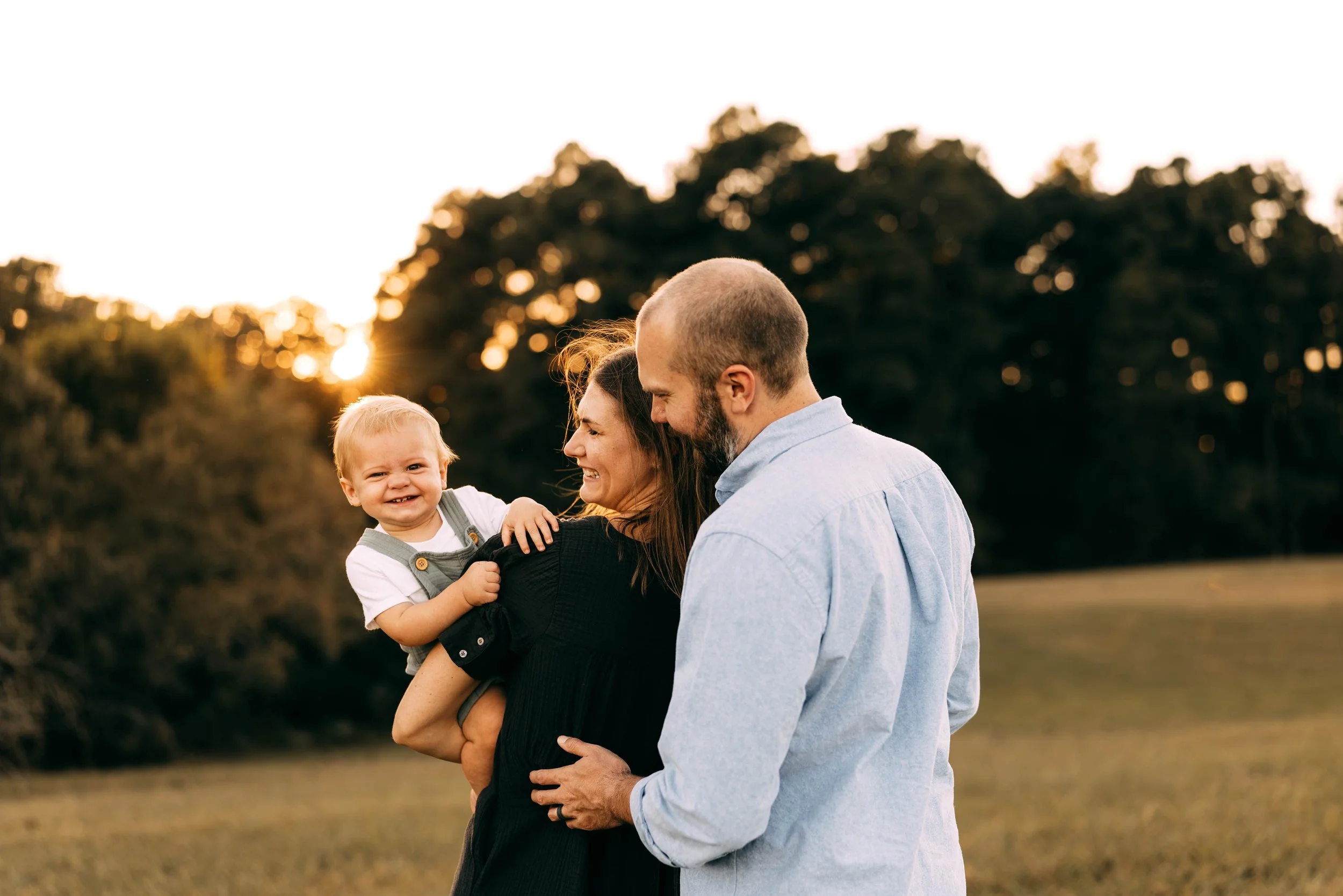 A happy family of three outdoors at sunset, with the mother holding a smiling young boy and the father standing close, all enjoying a joyful moment together.