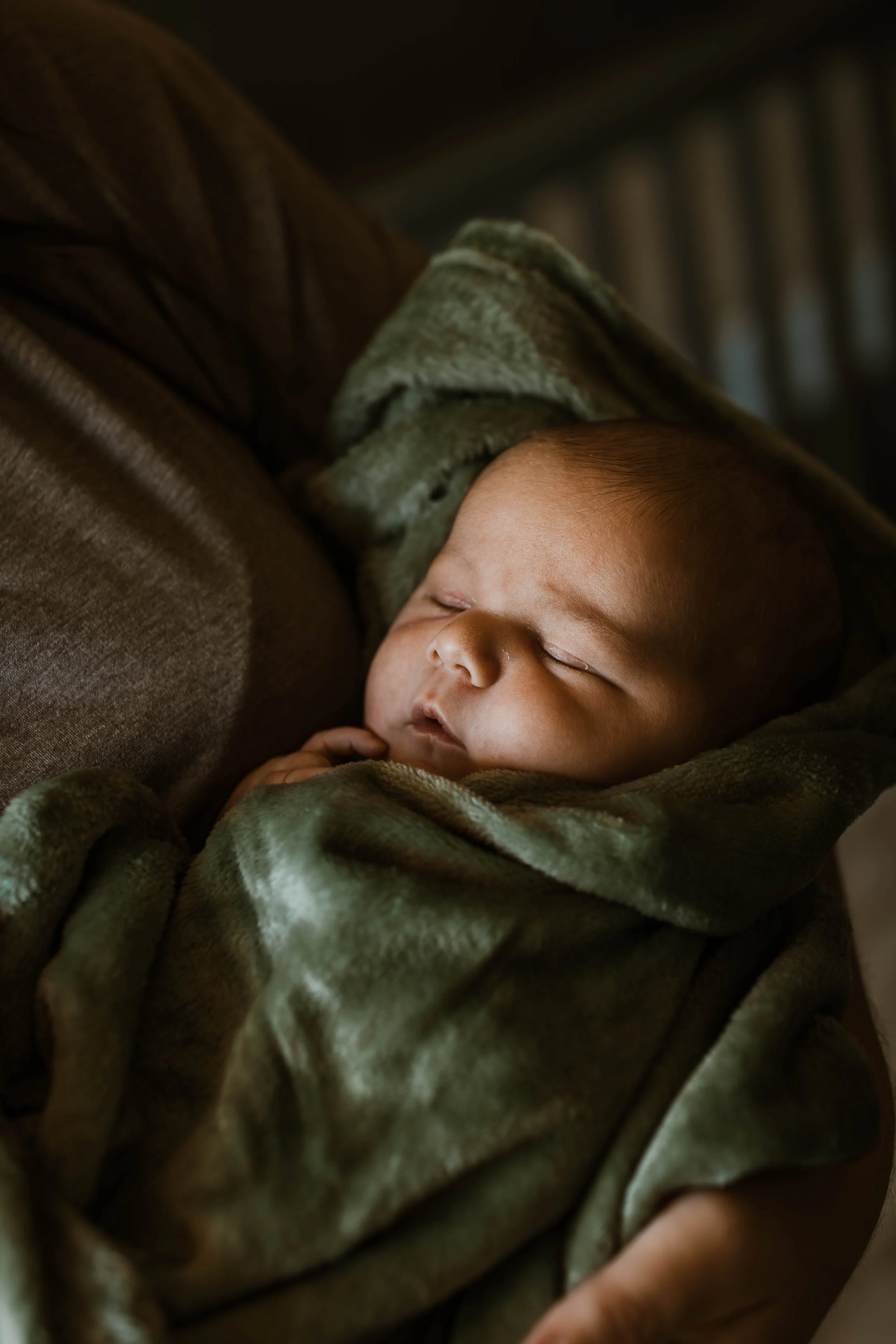 A sleeping baby wrapped in a green blanket, resting on a brown couch.