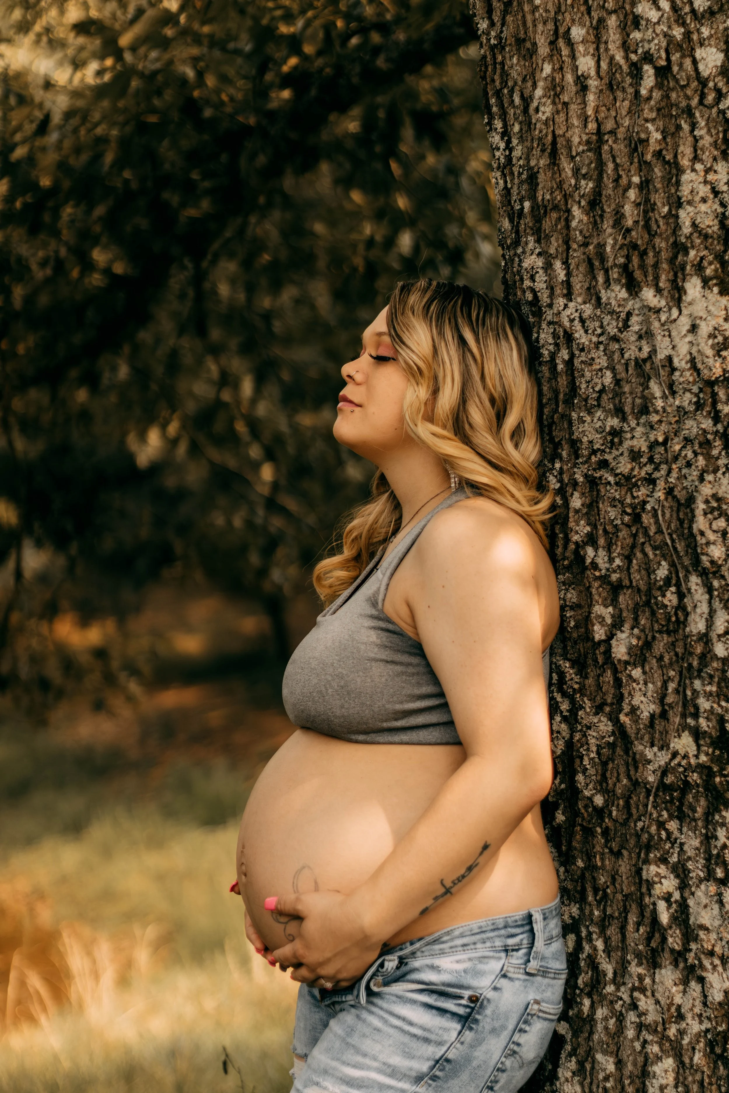 Pregnant woman in gray sports bra and denim shorts leaning against a large tree with closed eyes, surrounded by nature.