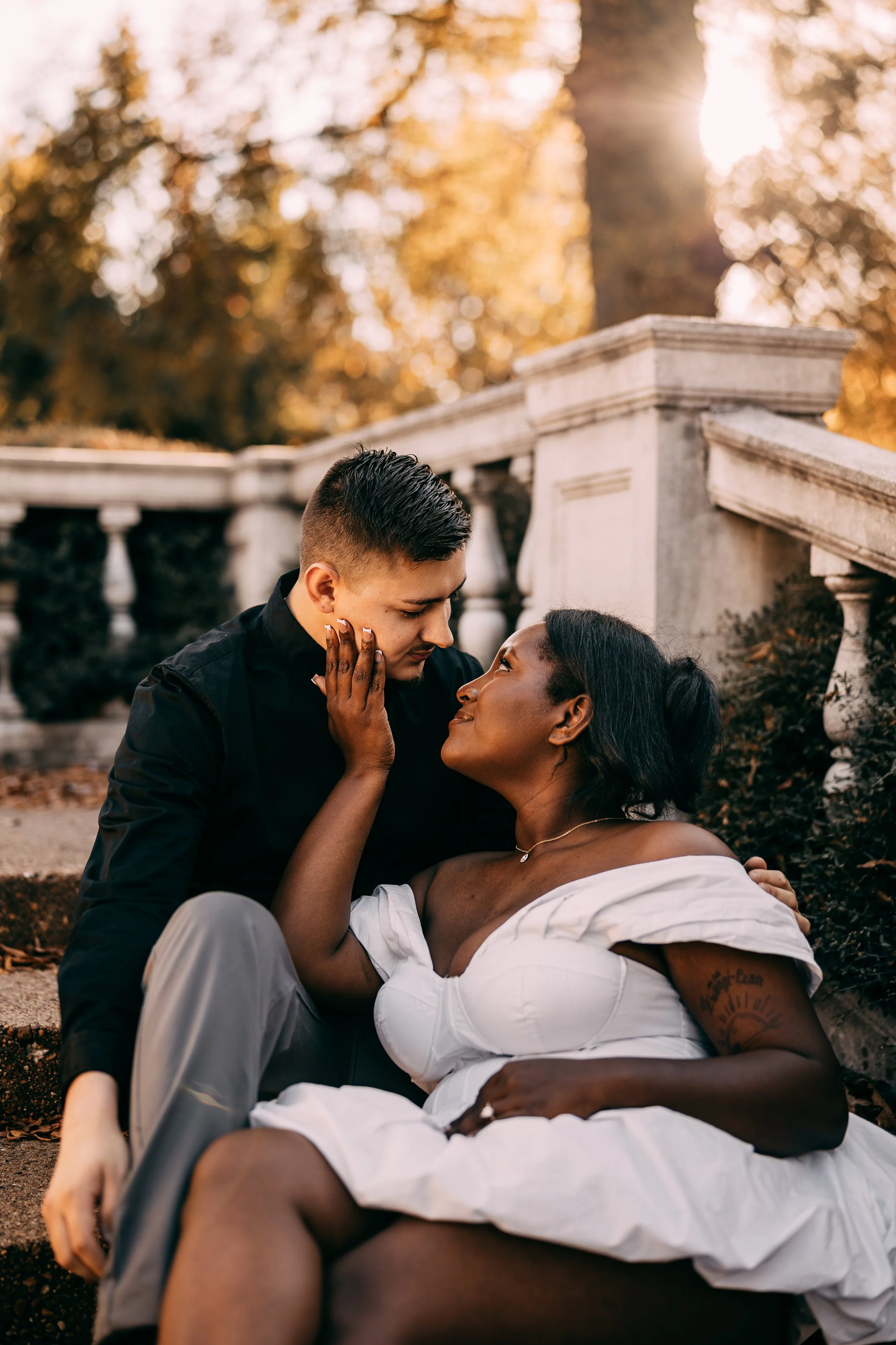 A couple sitting on outdoor stone stairs, gazing into each other's eyes, with autumn trees and sunlight in the background.