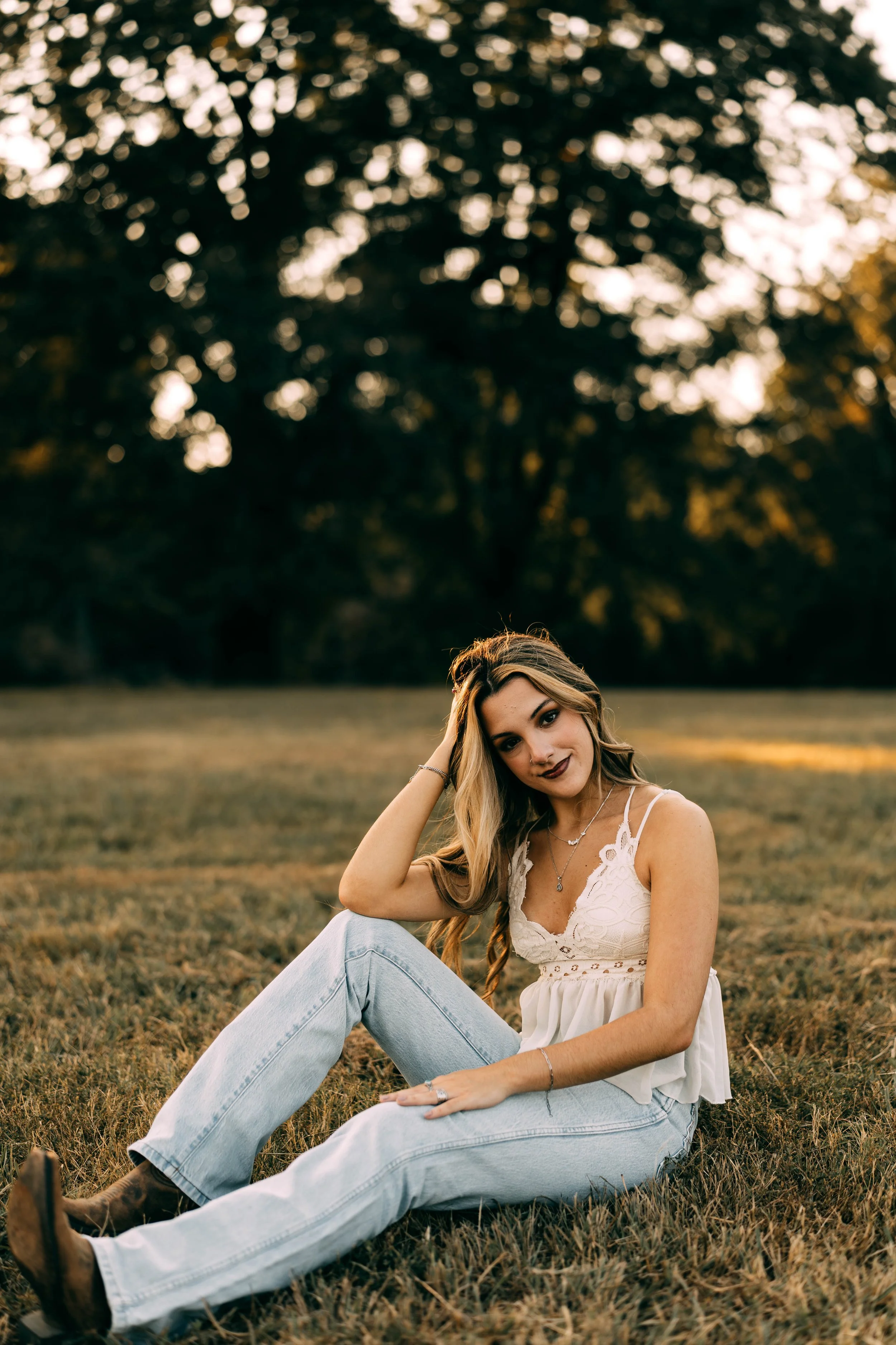 A young woman sitting on grass in a park during sunset, wearing a white lace top, light jeans, and boots, with long wavy hair, smiling gently at the camera.