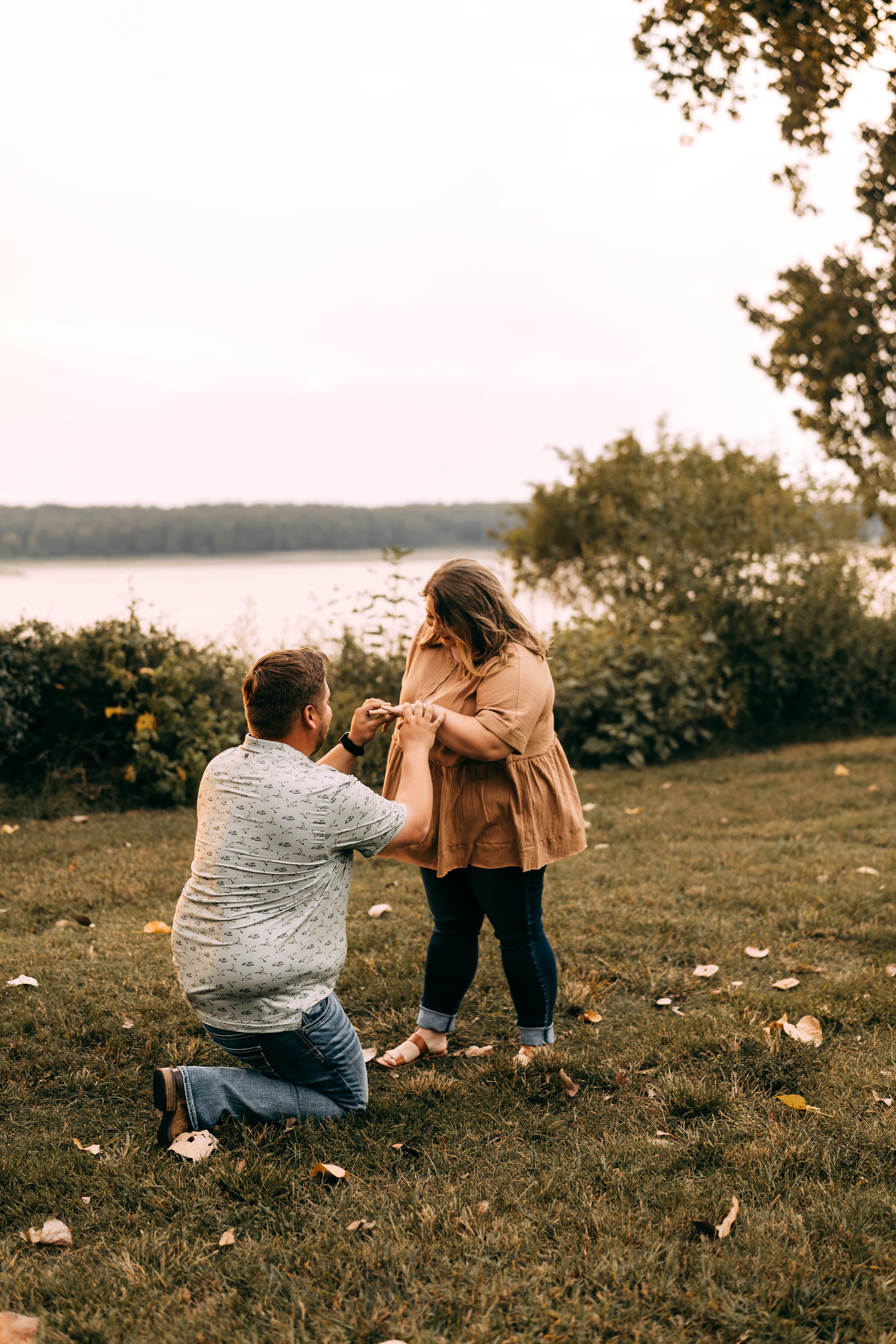 A man proposing to a woman outdoors during sunset, with trees and a body of water in the background.