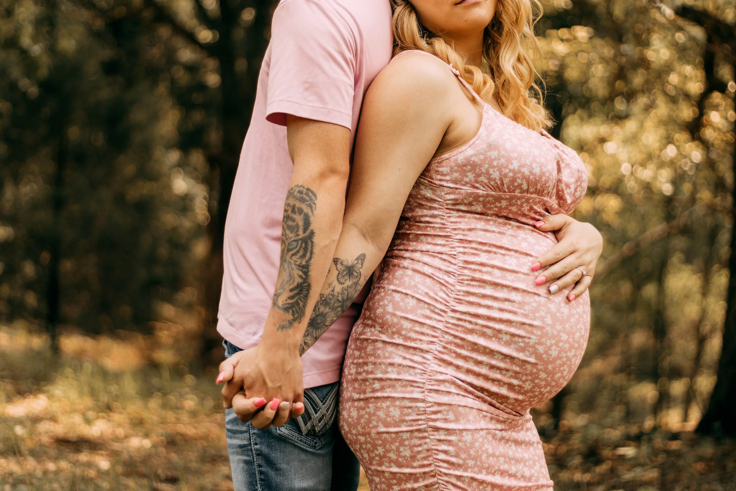 A pregnant woman in a pink floral dress holding her belly, with a partner behind her, holding hands in a wooded outdoor park setting.