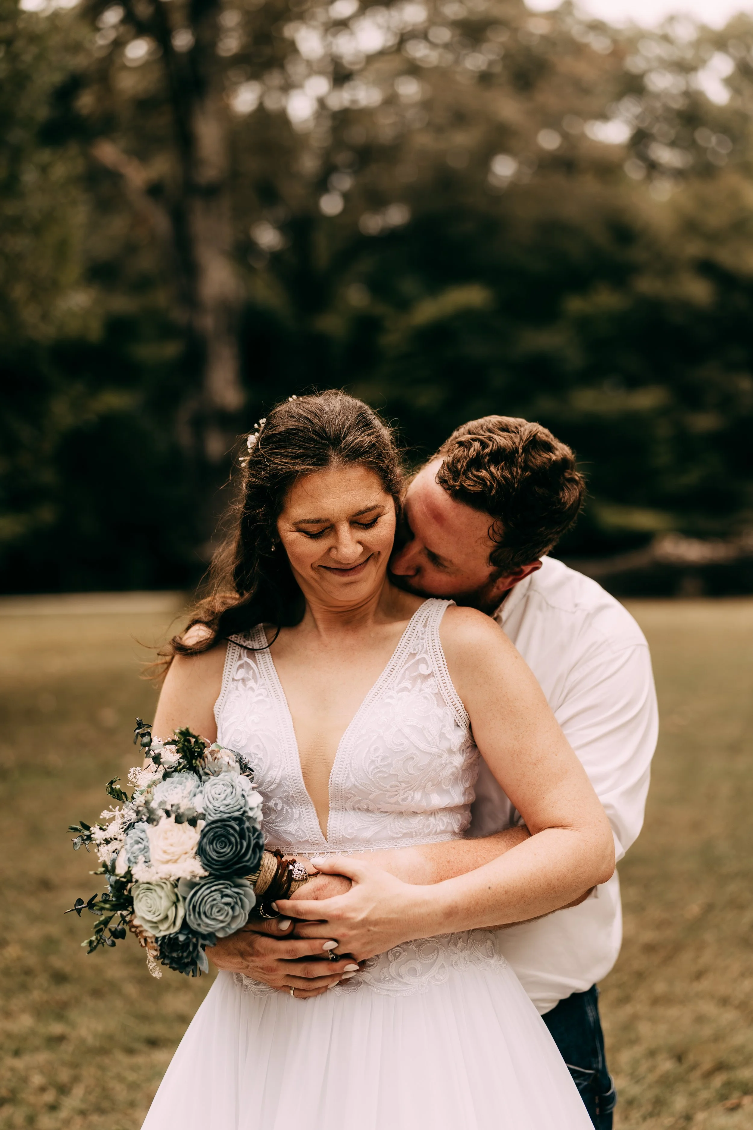 A couple in wedding attire embracing outdoors, the woman holding a bouquet of flowers, on a grassy area with trees in the background.