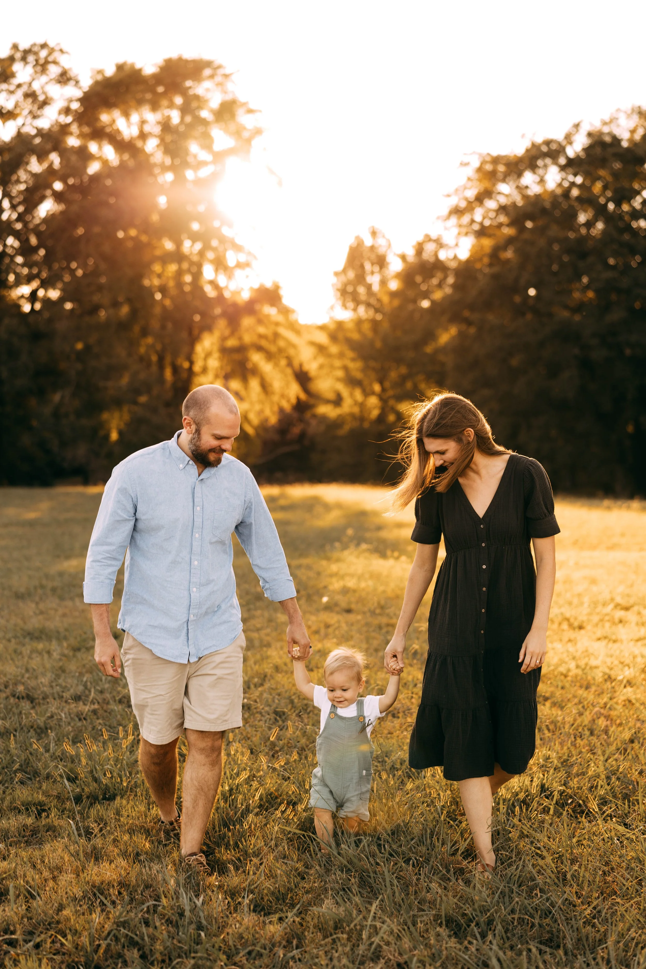A family of three, including a father, mother, and toddler, walking hand in hand in a grassy field during sunset. The sun is shining through trees in the background, creating a warm, golden glow.