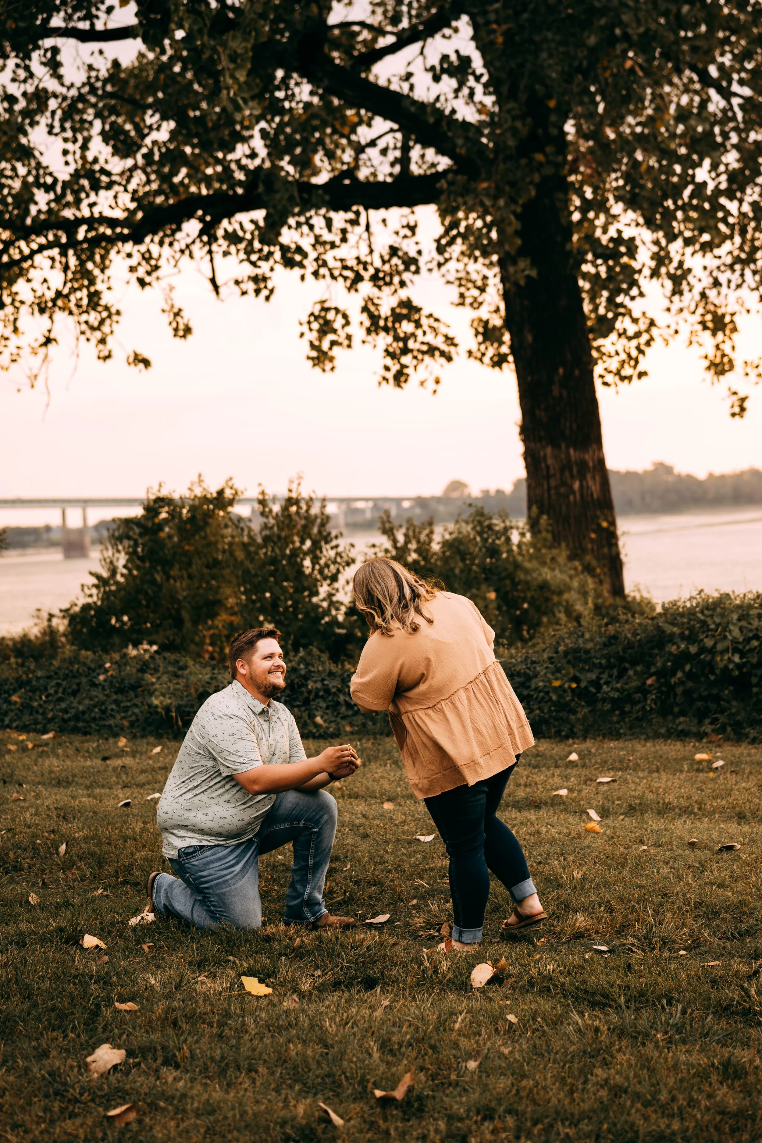 A man on one knee proposing to a woman in a park beside a river, with a large tree overhead and a bridge in the background during sunset.