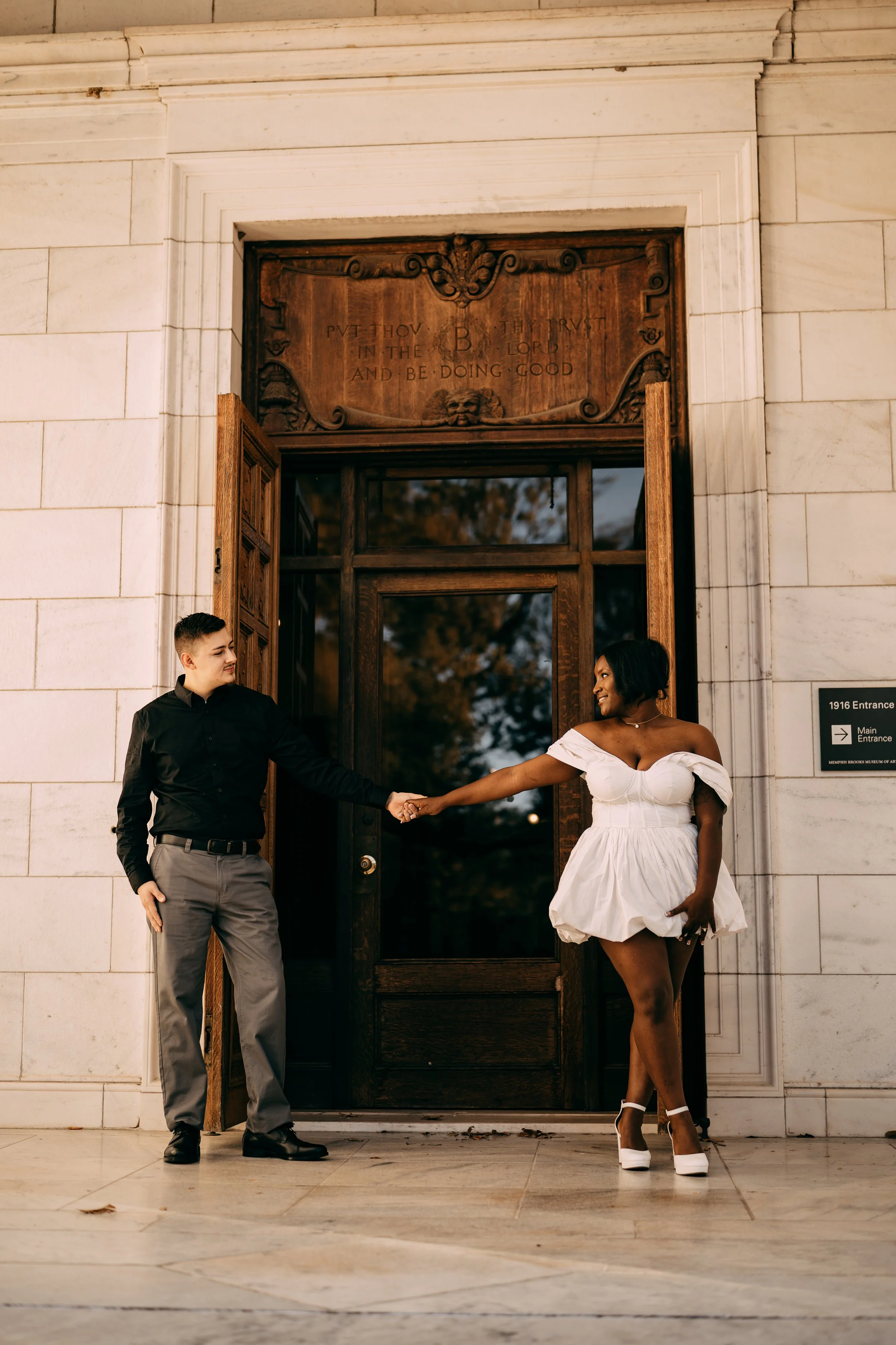 A couple is standing in front of an entrance door, holding hands and smiling. The man is dressed in a black shirt and gray pants, and the woman is wearing a white off-the-shoulder dress with white high heels.