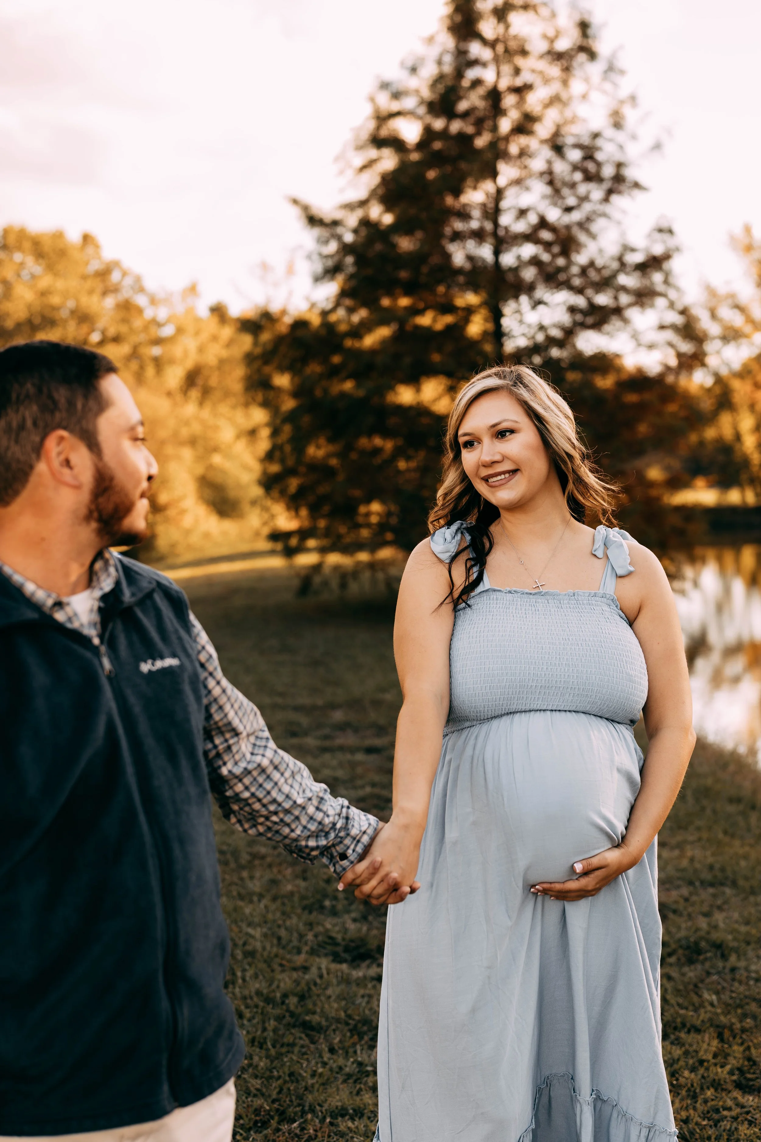 A pregnant woman in a light blue dress holds her belly with one hand and holds hands with a man dressed in a plaid shirt and vest near a lake during sunset.