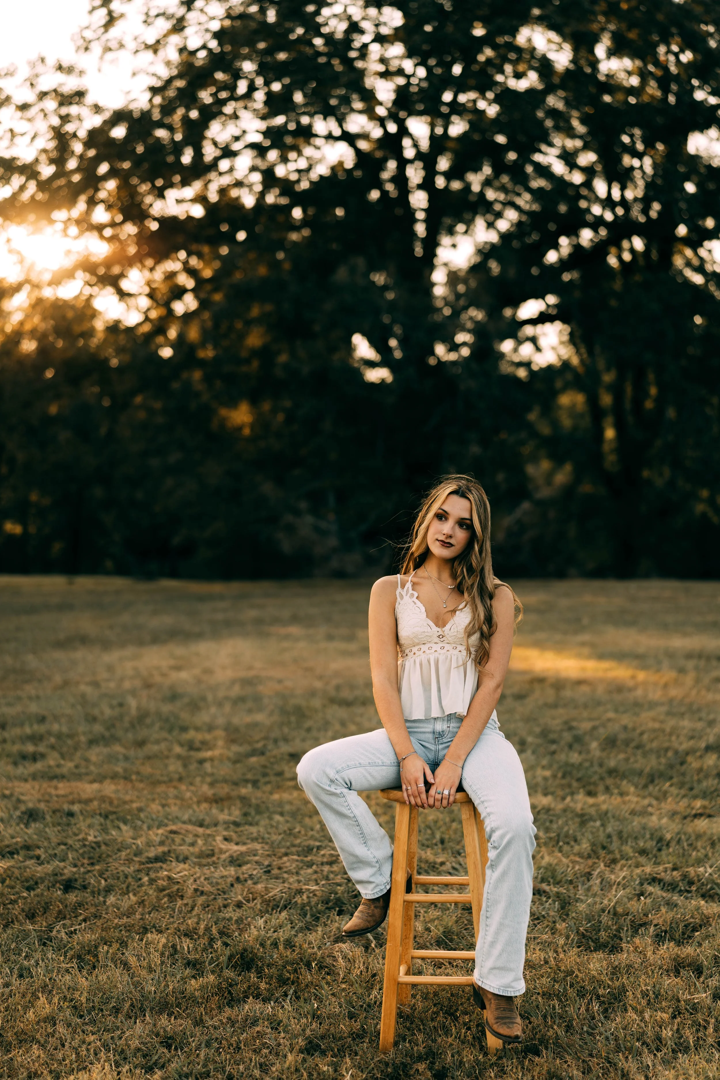 A young woman sitting on a wooden stool outdoors in a grassy field during sunset, with trees in the background.
