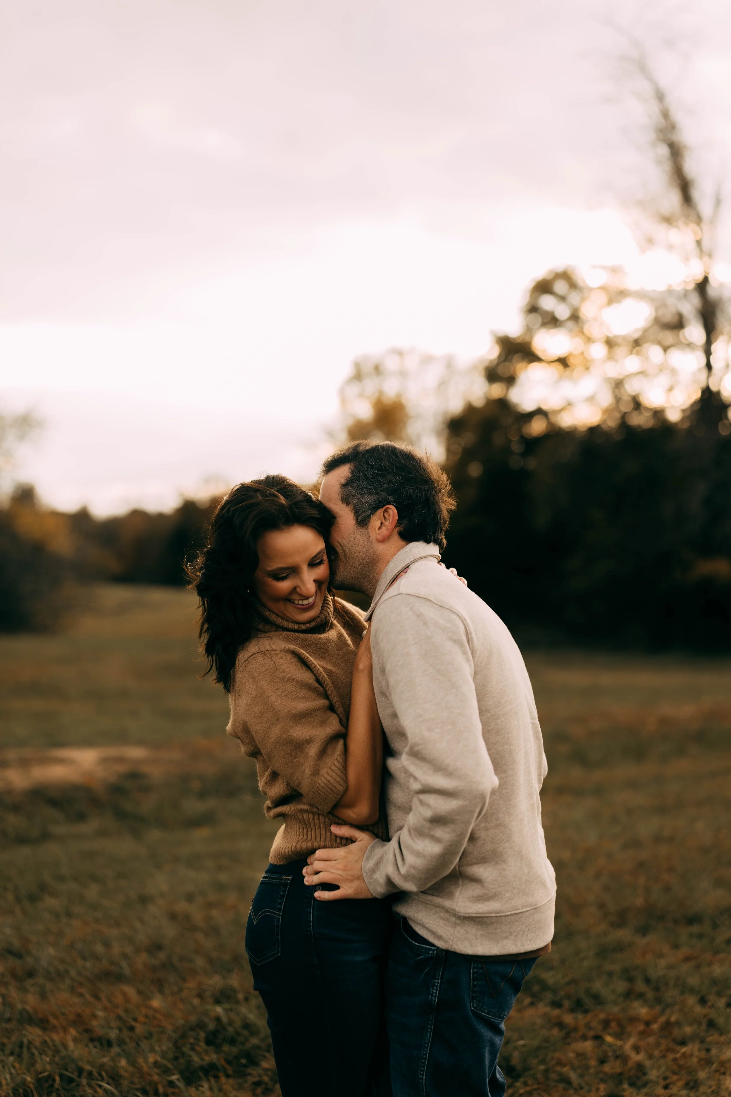 A couple smiling and hugging in a park during sunset, with trees in the background.