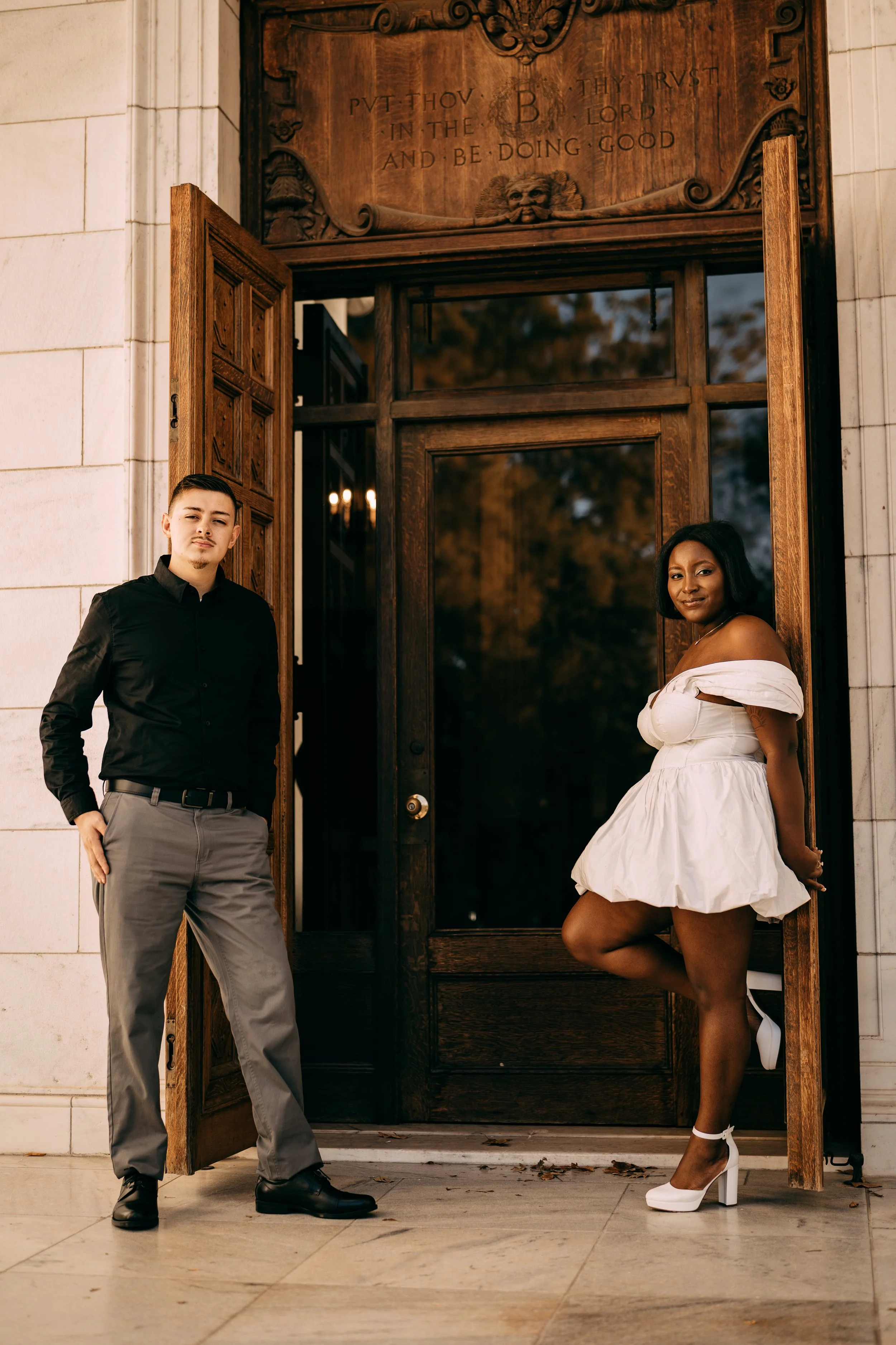 A young man and woman stand near the entrance of a building with ornate wooden doors. The man is dressed in a black shirt and gray pants, and the woman is wearing a white off-the-shoulder dress and white high heels. The doors are open, revealing a re