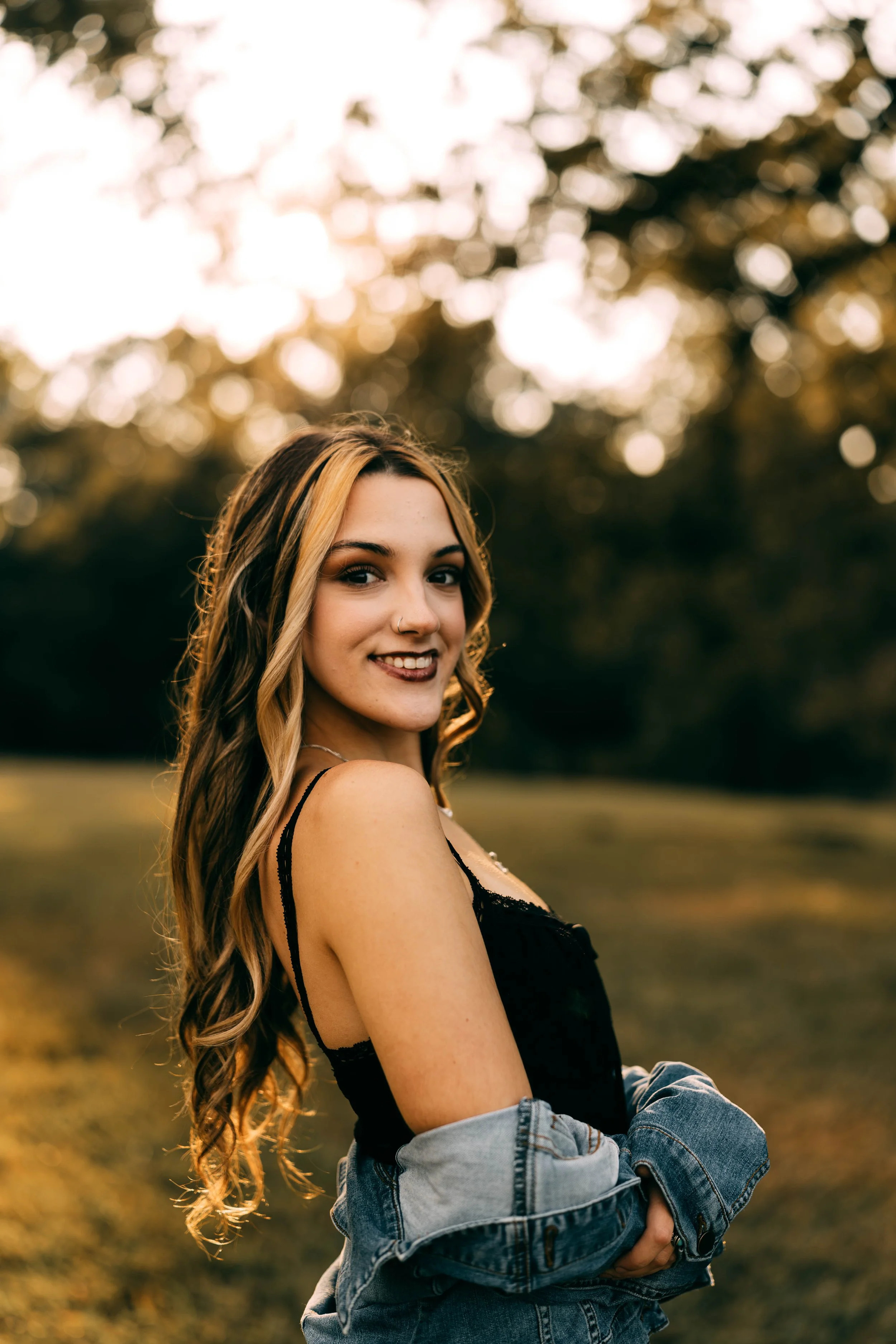 A young woman with long wavy hair, smiling, wearing a black top and a denim jacket draped over her shoulders, standing outdoors in a park at sunset.