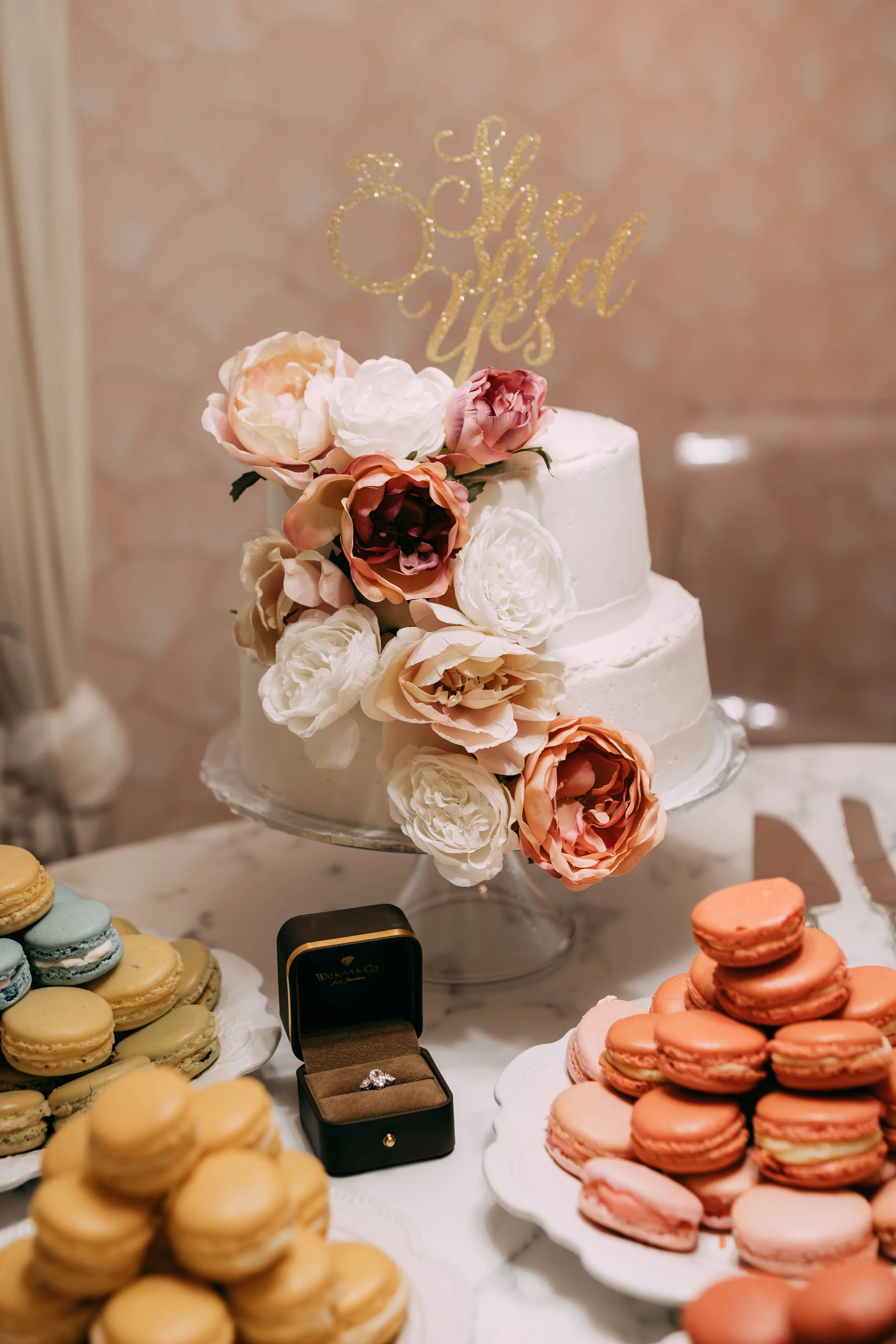 Wedding cake decorated with a cascade of pink, white, and peach flowers, topped with a gold 'she said yes' cake topper, surrounded by macarons and a diamond ring in a black box.